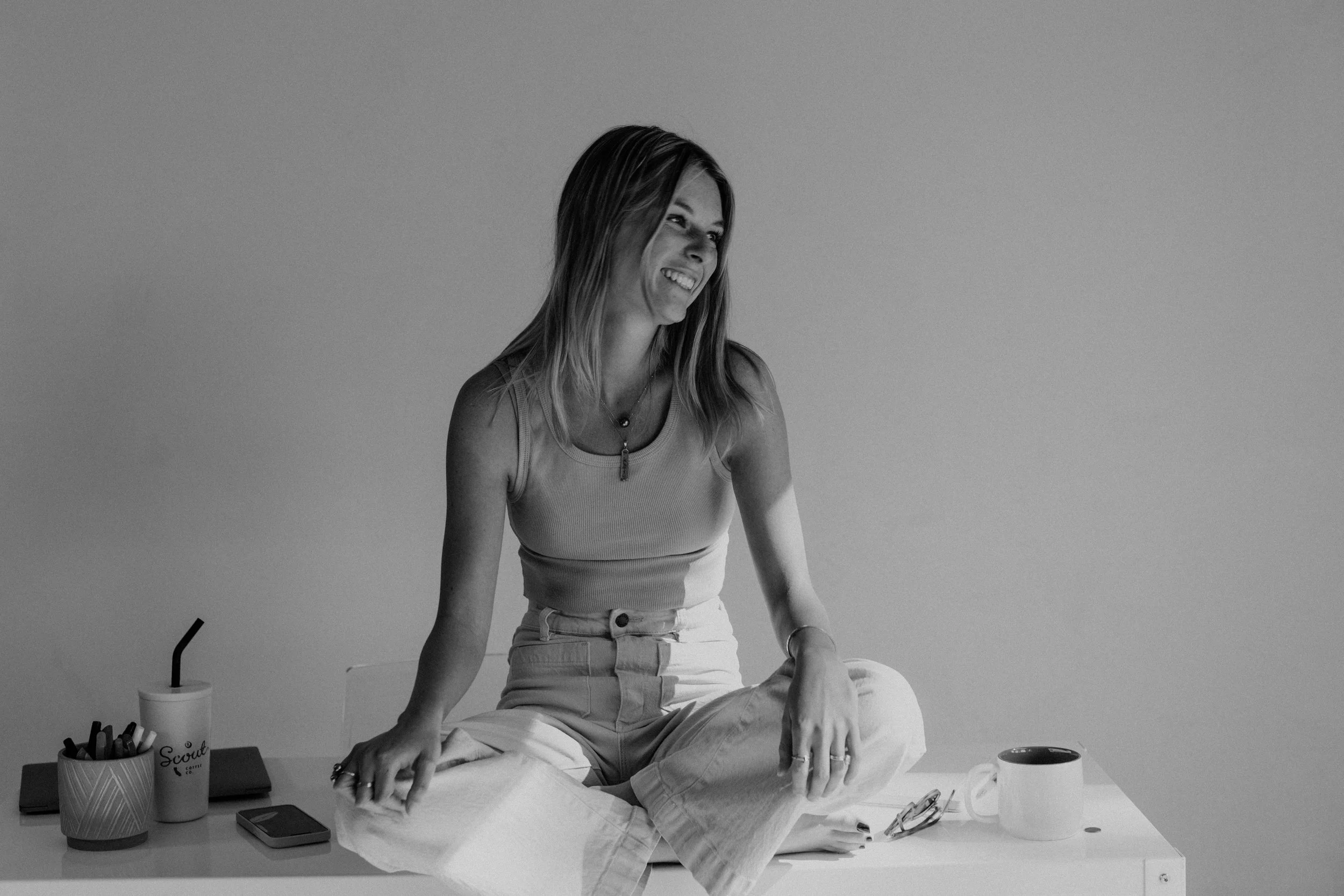 A woman sitting cross-legged on a desk, smiling and looking to her right, with a coffee mug, glasses, a pen holder, and a phone on the desk.