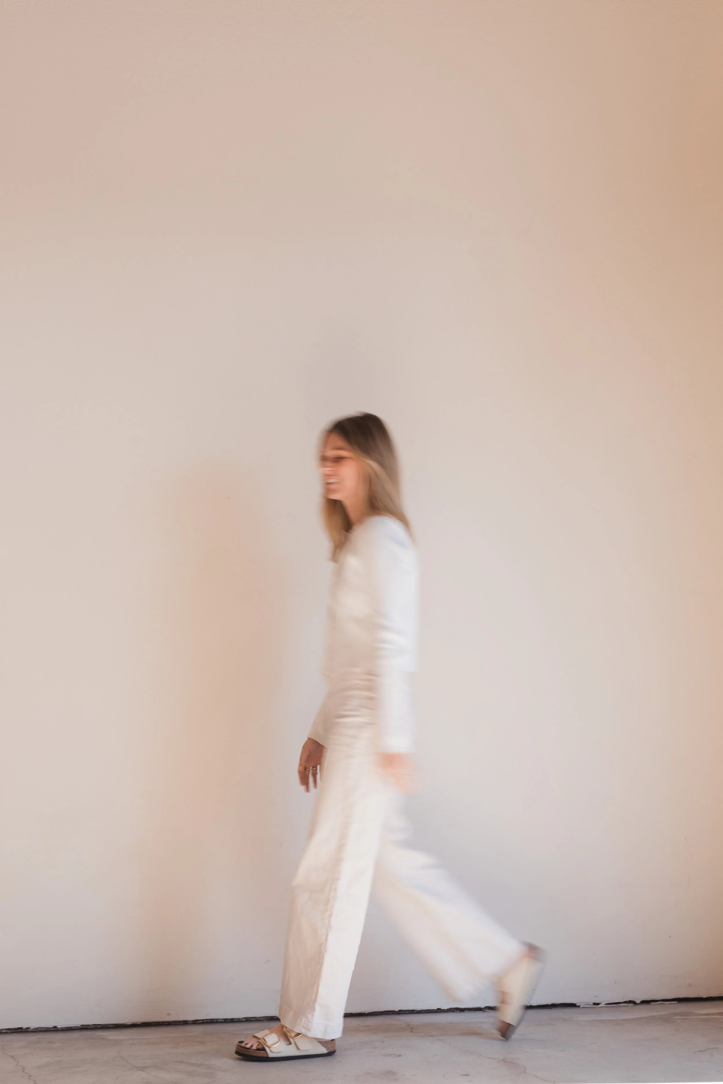 A woman in white pants and blazer walking, smiling, against a plain light-colored wall.