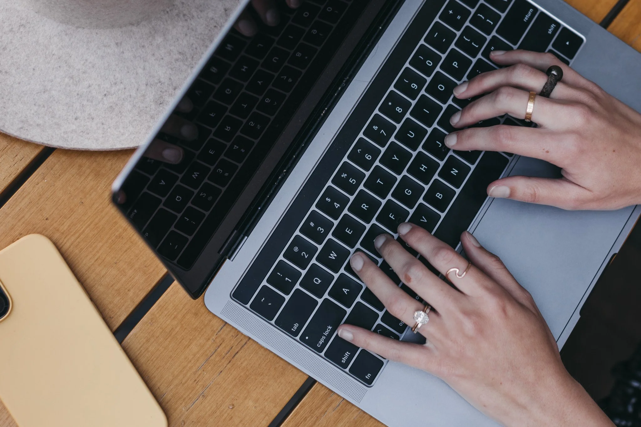 Person typing on a laptop keyboard with rings on fingers, on a wooden table next to a yellow phone and a beige hat doing bookkeeping and tax prep on the Central Coast, CA.
