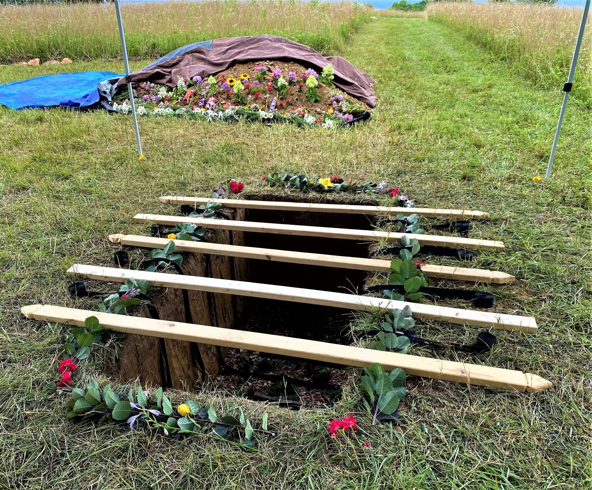 Flowers line a grave prepared for a natural burial. Equally-spaced boards cover the grave, with prairie in the background.