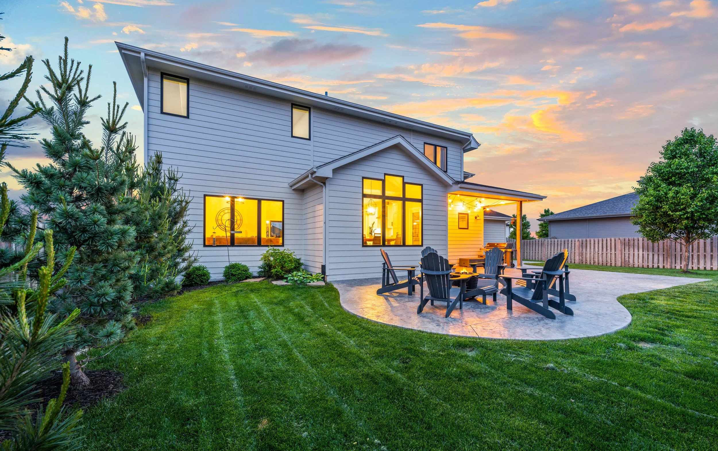 Twilight exterior photograph of a residential home with a stamped concrete patio, Adirondack seating around a fire pit, manicured lawn, and warm interior lighting against a dramatic sunset sky.