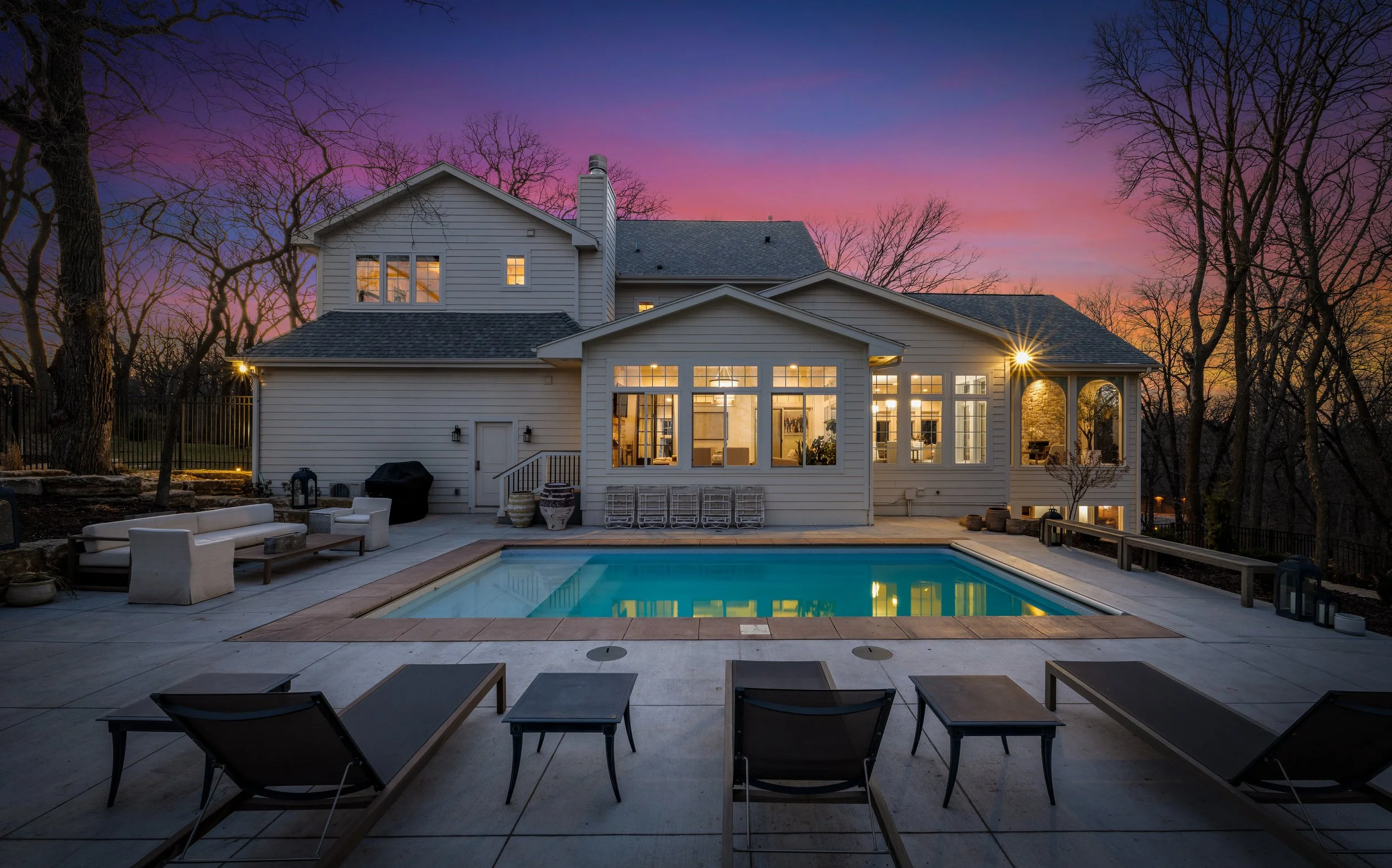 Twilight exterior photograph of a residential property with a pool, outdoor seating, and warm interior lighting glowing through large windows against a pink and purple sunset sky.