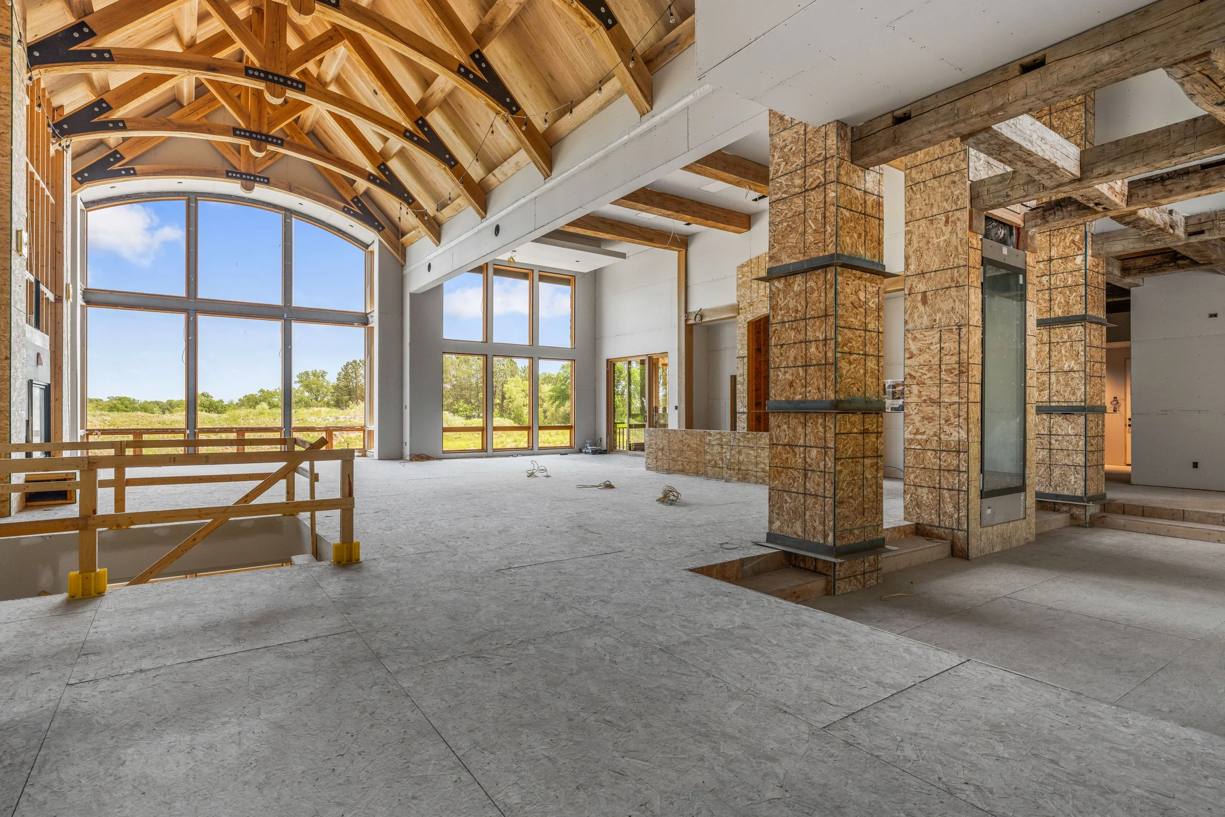 Framed great room with vaulted timber trusses and arched window during construction, drywall not yet installed
