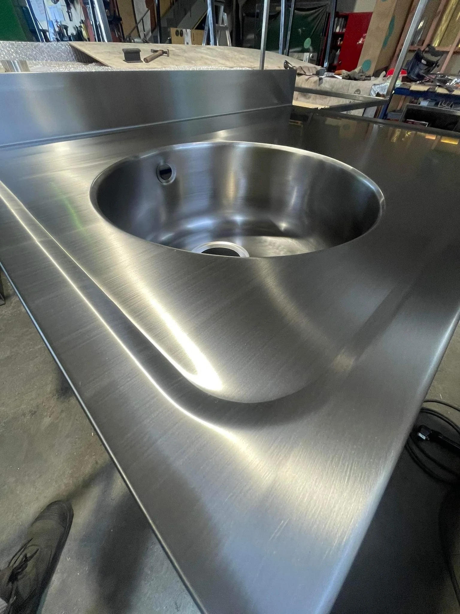 A stainless steel sink and countertop area in a workshop or industrial setting, with tools and equipment visible in the background.