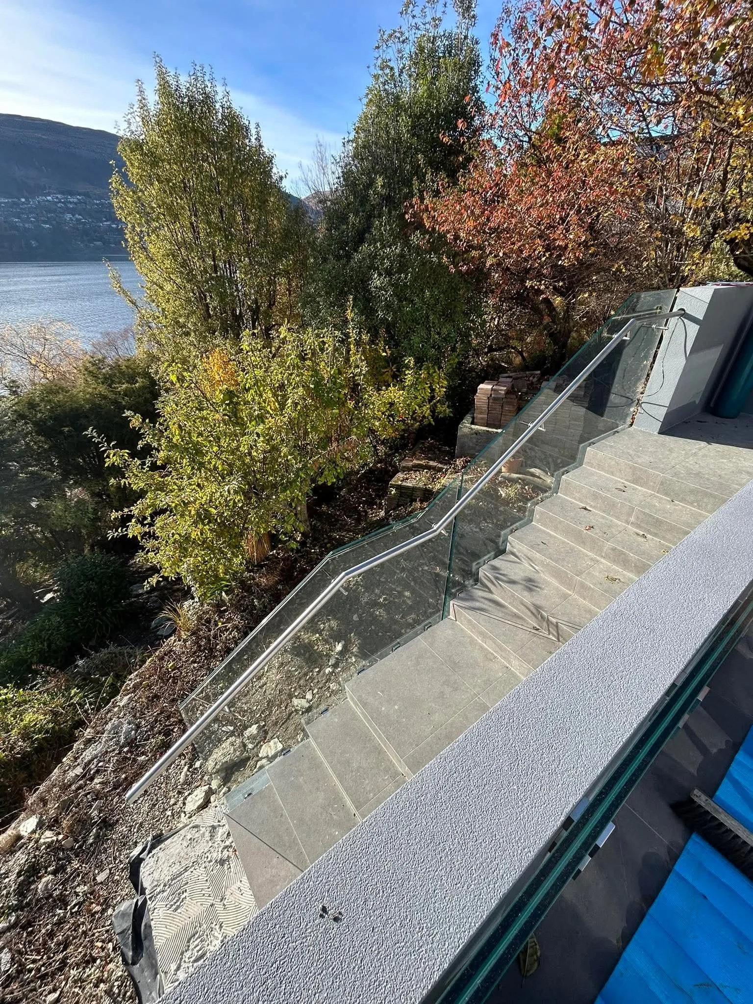 Outdoor scene showing a staircase with glass railing overlooking trees with green and red leaves, a body of water, and mountains in the background on a sunny day.