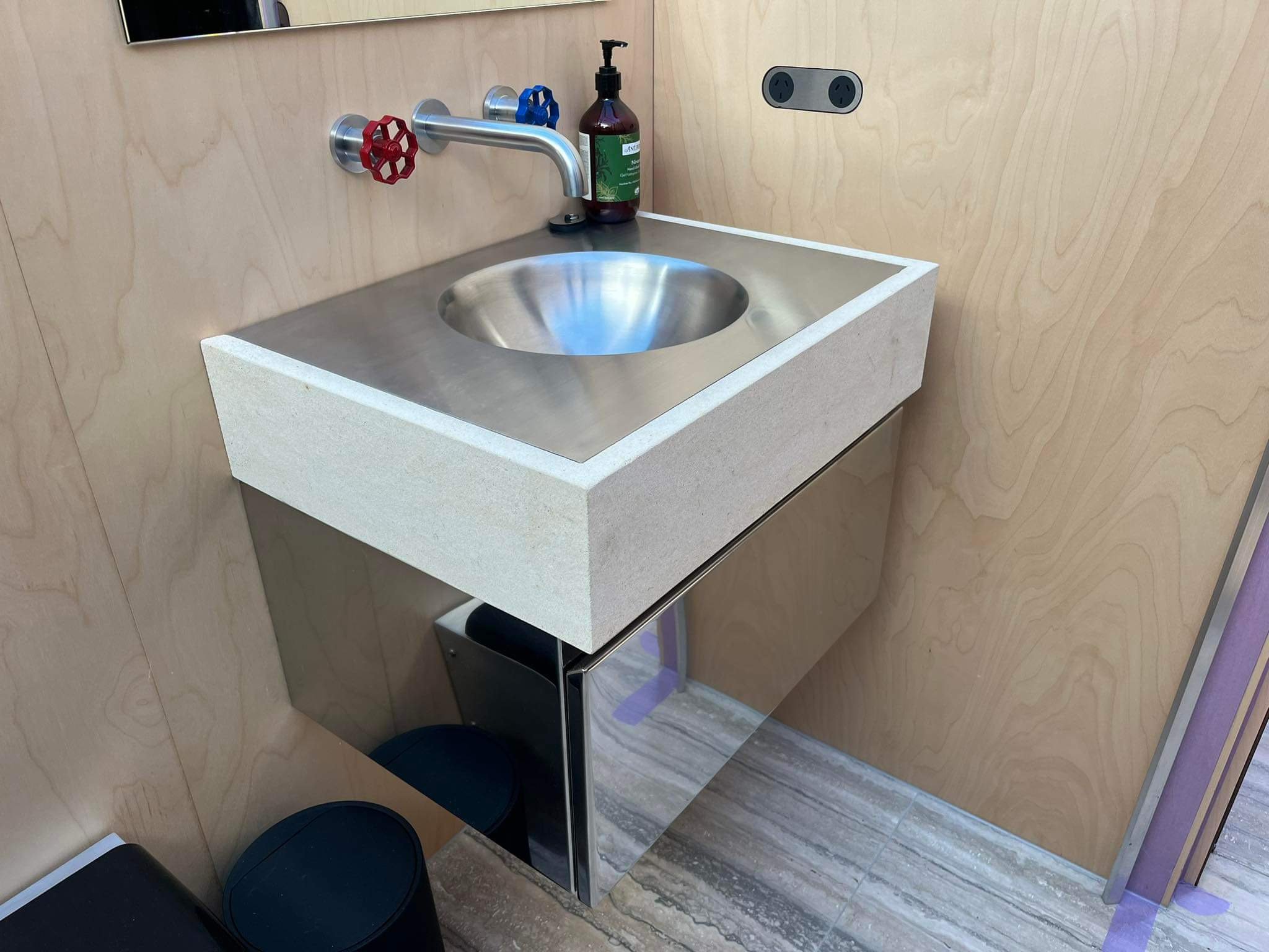 A small stainless steel sink with a wall-mounted faucet that has red and blue handles, a soap dispenser, and a bottle of hand soap on the counter. The scene appears to be in a public restroom or similar facility.