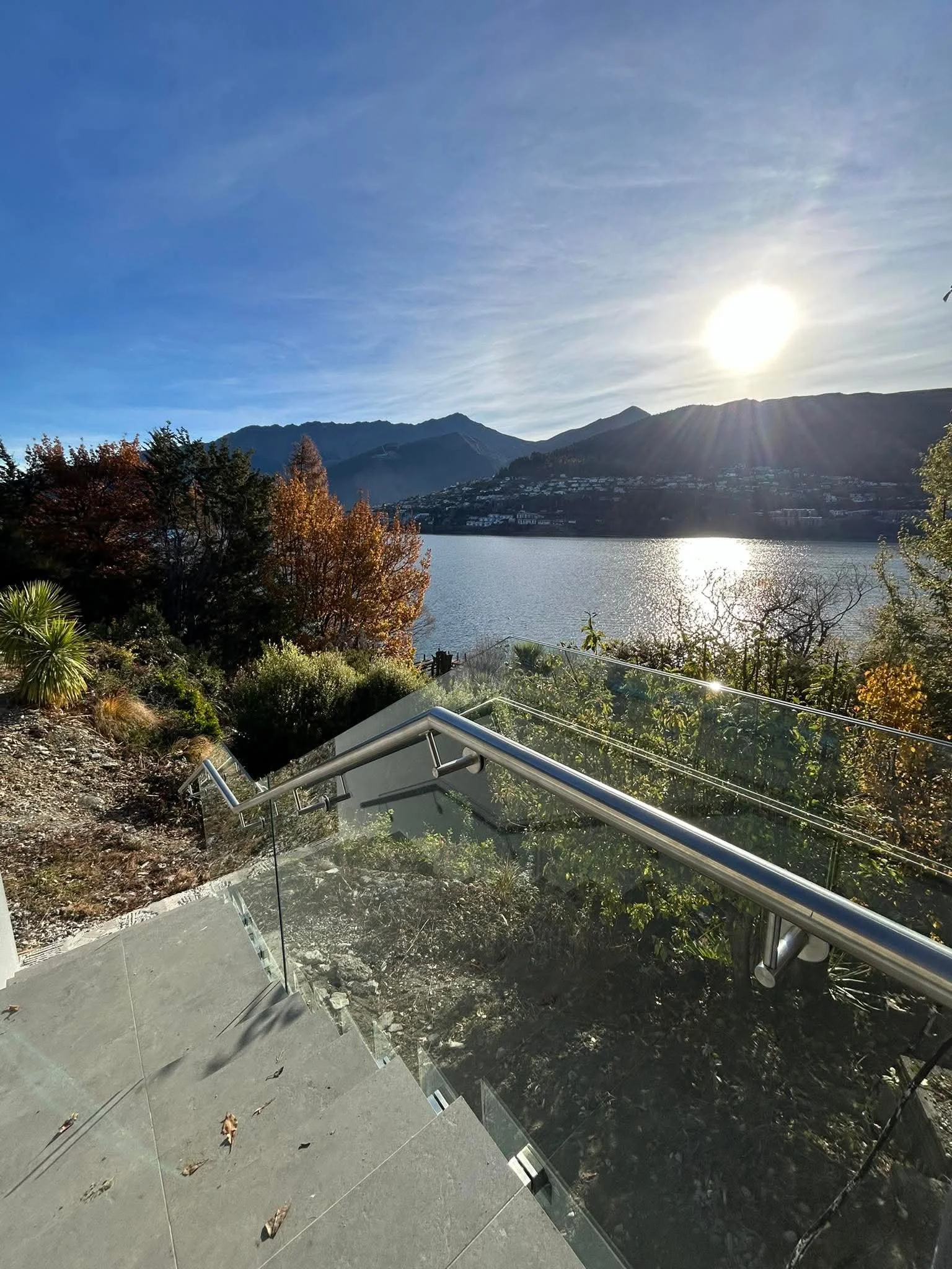 Scenic view of a lake surrounded by mountains with the sun shining, trees with autumn foliage, and a glass railing in the foreground.