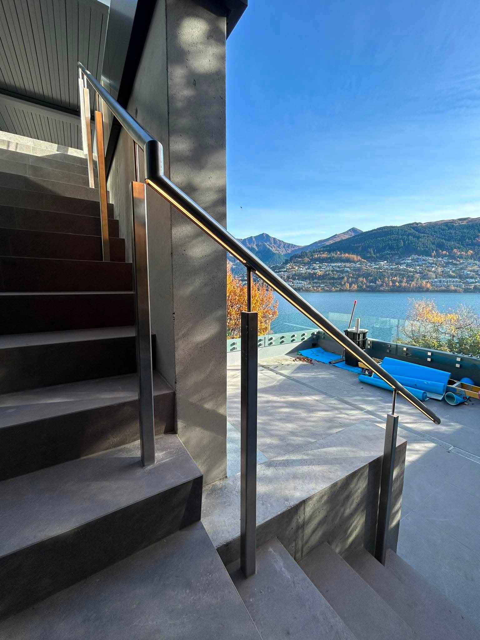 Modern staircase with metal handrail and concrete steps, overlooking a scenic view of mountains and a body of water with blue sky in the background.