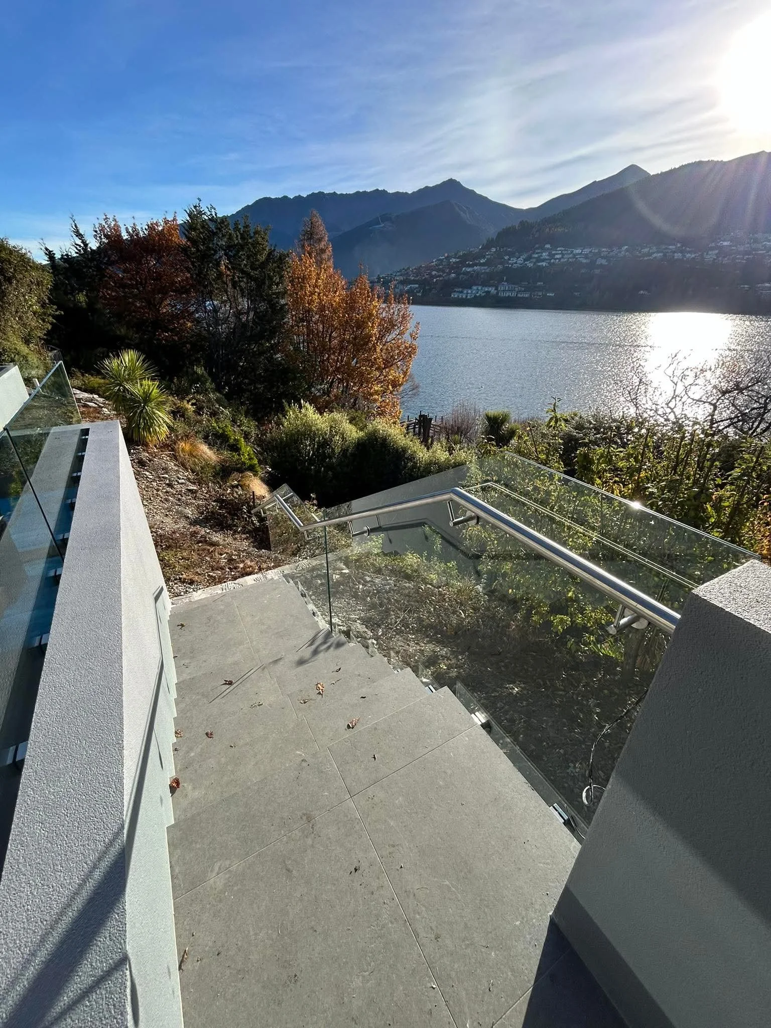 Concrete staircase with glass railing leading down to a garden area with bushes and trees, overlooking a large body of water with mountains in the background under a partly cloudy sky.
