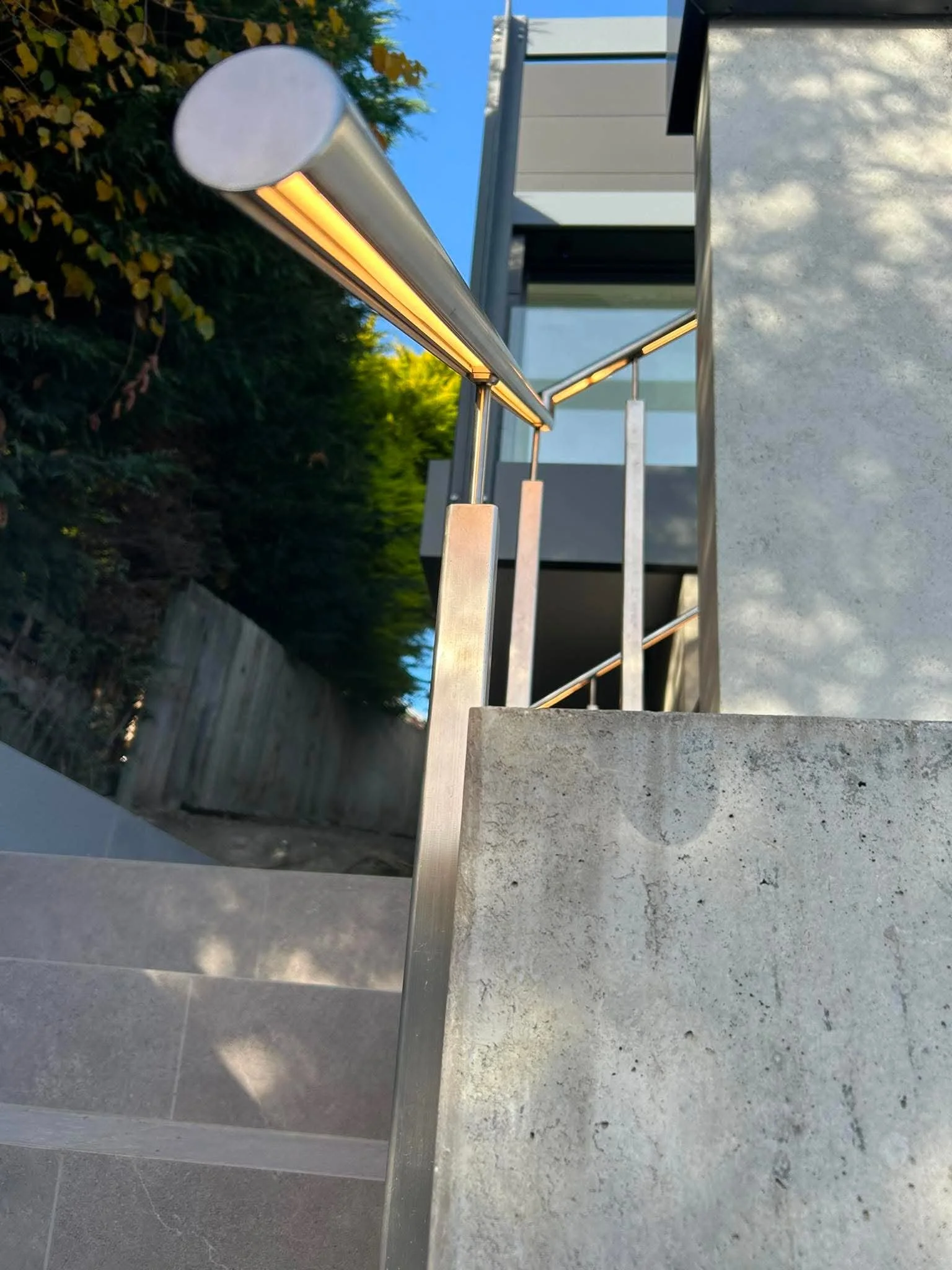 Close-up of a modern outdoor metal handrail on a concrete staircase, with greenery and blue sky in the background.
