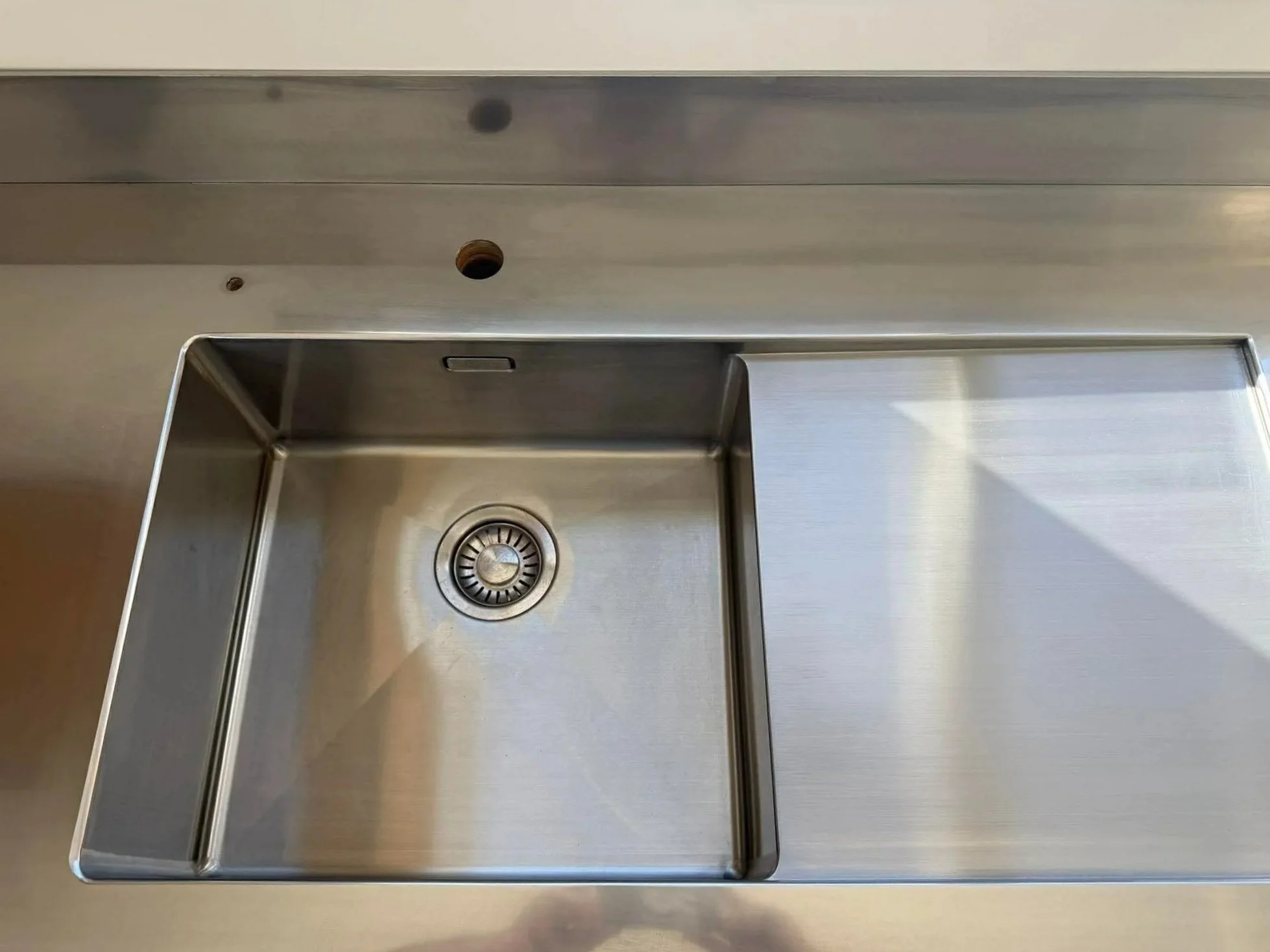 A stainless steel kitchen sink with a single basin and a drain at the bottom, set into a light-colored countertop.