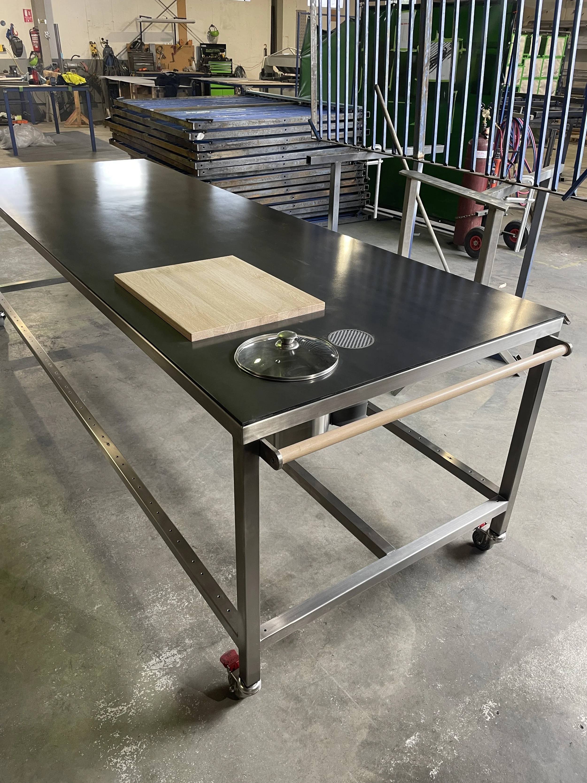 A stainless steel worktable with a black top surface, a wooden cutting board, a glass lid, and a drain on a concrete workshop floor with various tools and equipment in the background.