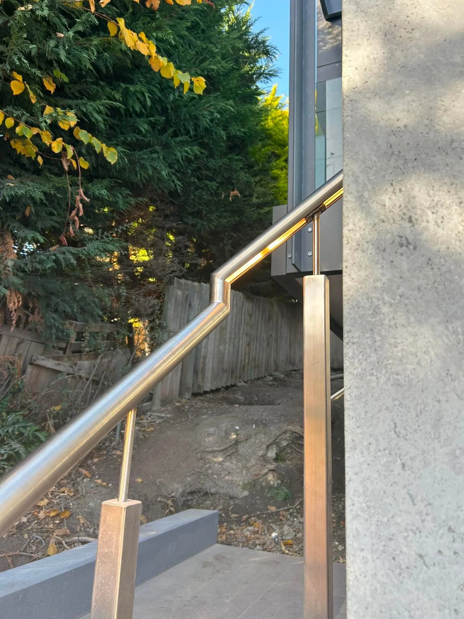 The image features a modern outdoor staircase with a metal handrail, next to a concrete wall, with a lush green tree and a blue sky in the background.