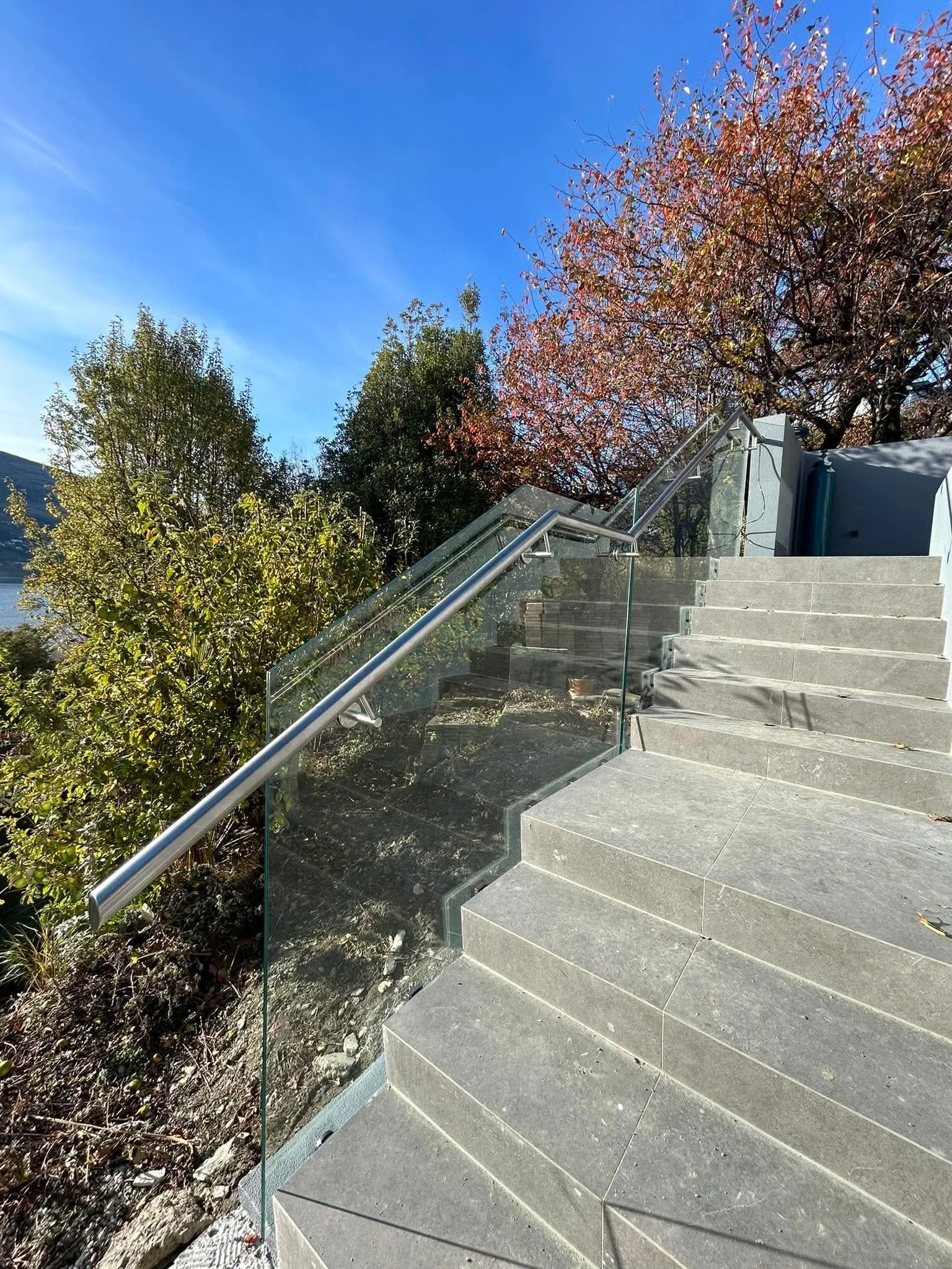 Concrete staircase with a glass and metal railing, surrounded by trees with green and orange leaves, under a blue sky.