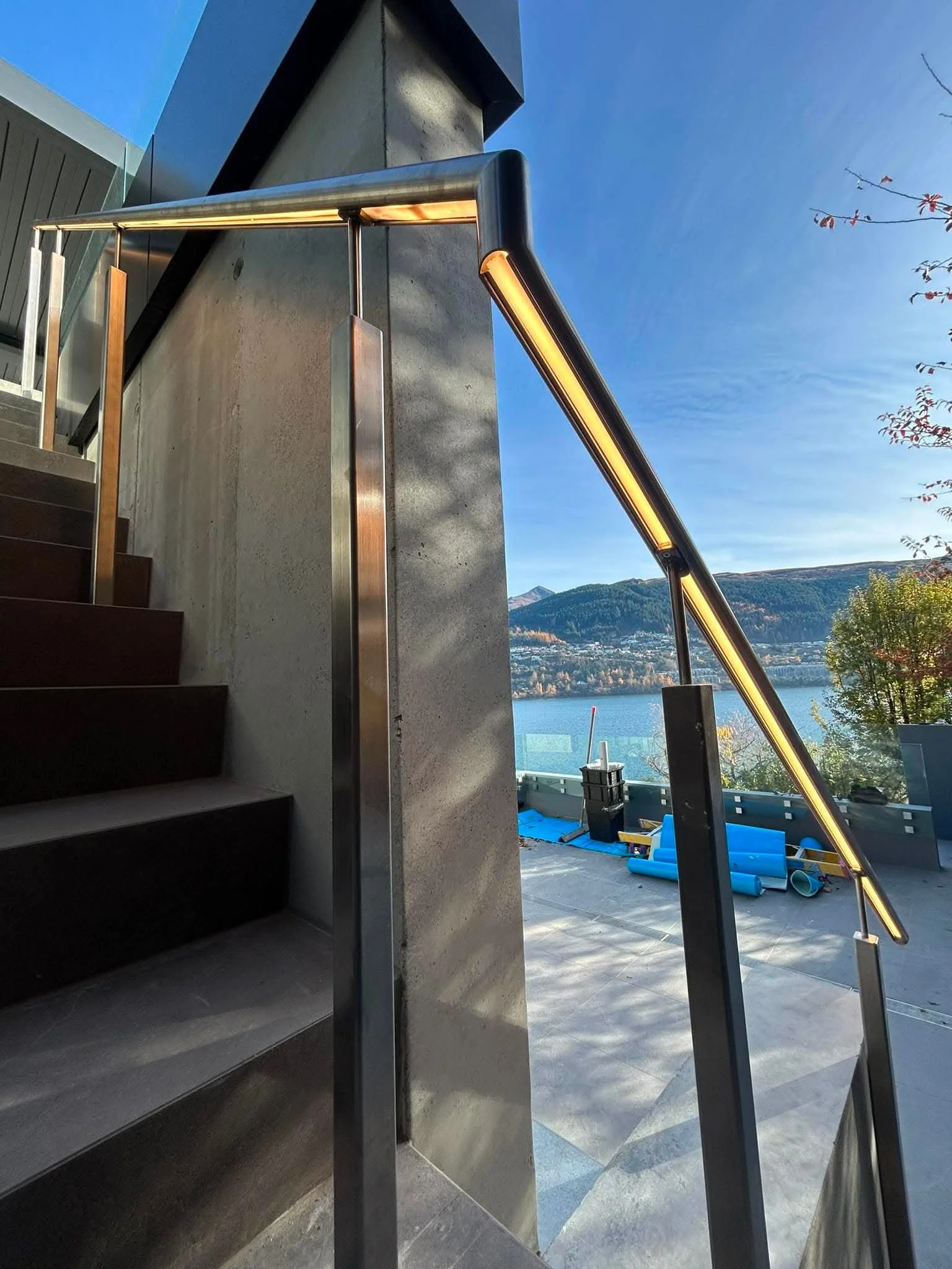 A modern staircase with dark wooden steps and a metal handrail, leading up to an open outdoor patio with a view of a body of water, mountains, and trees, under a clear blue sky.