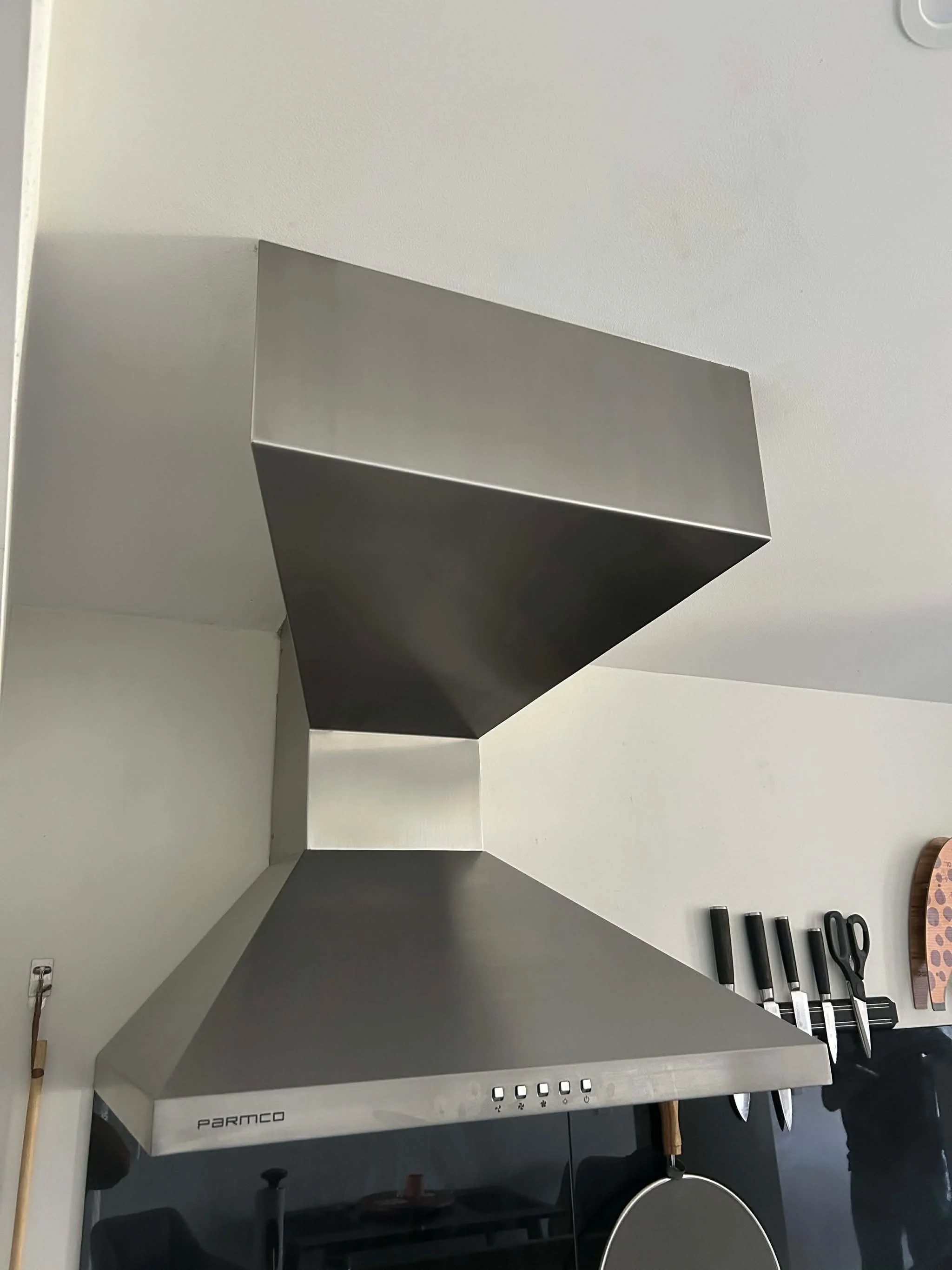 Commercial kitchen hood with the brand name 'Parmco' on it, four black knives with white handles on a magnetic strip on the wall, and part of a round frying pan hanging from a hook.