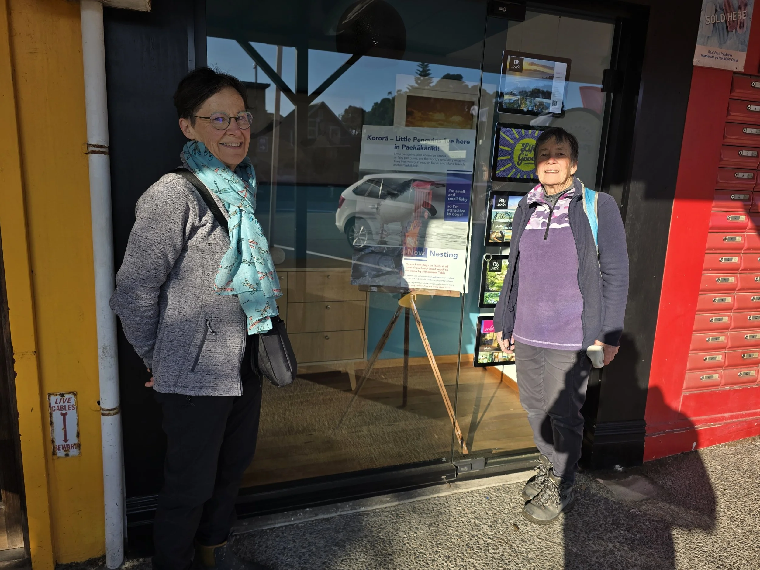 Kapiti Kororā Project volunteers Sue and Glenda in front of kororā advocacy signage in the Paekākāriki village