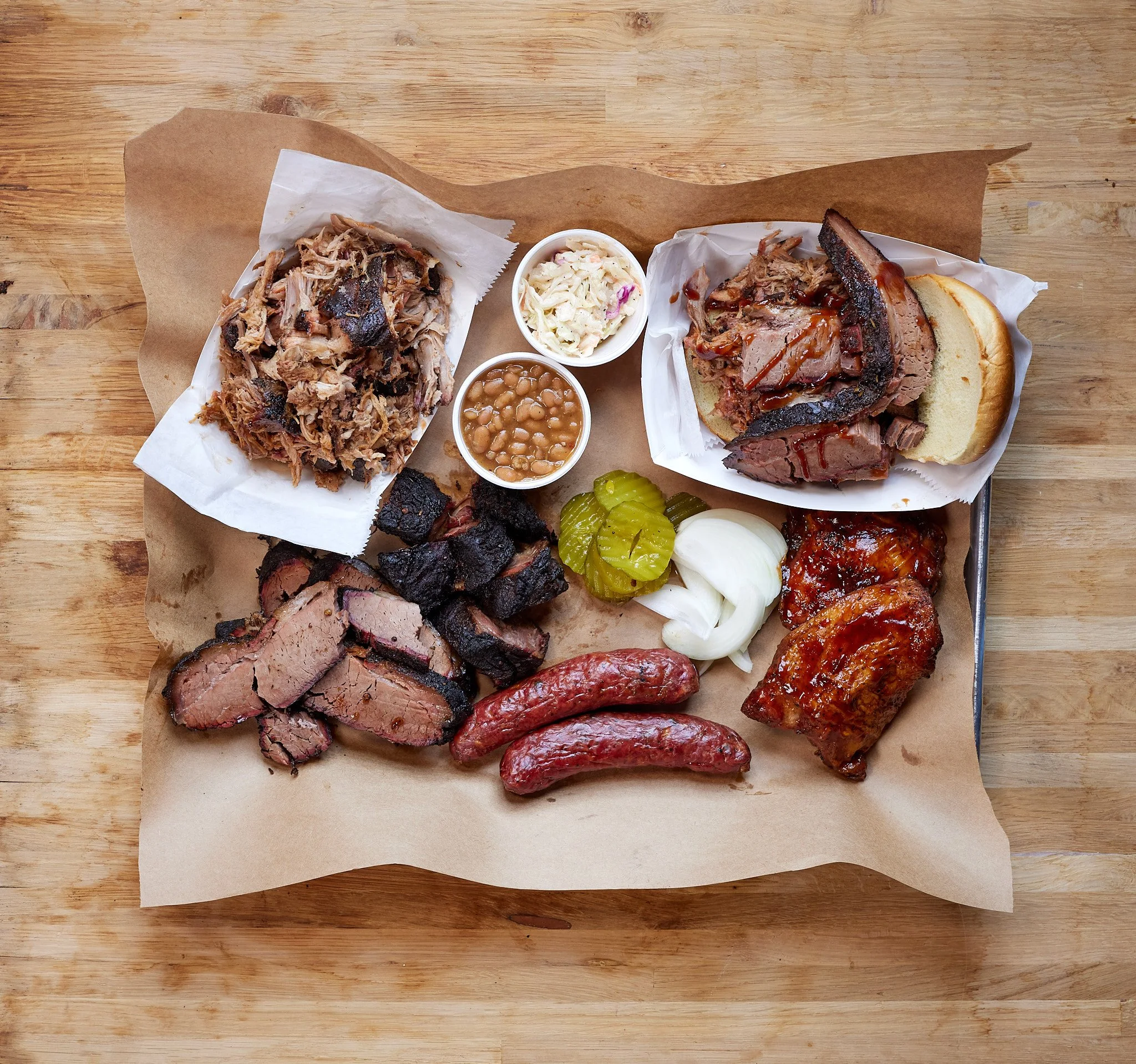 A barbecue platter with pulled pork, sliced brisket, sausages, ribs, pickles, onions, baked beans, coleslaw, and slices of bread on a wooden table.