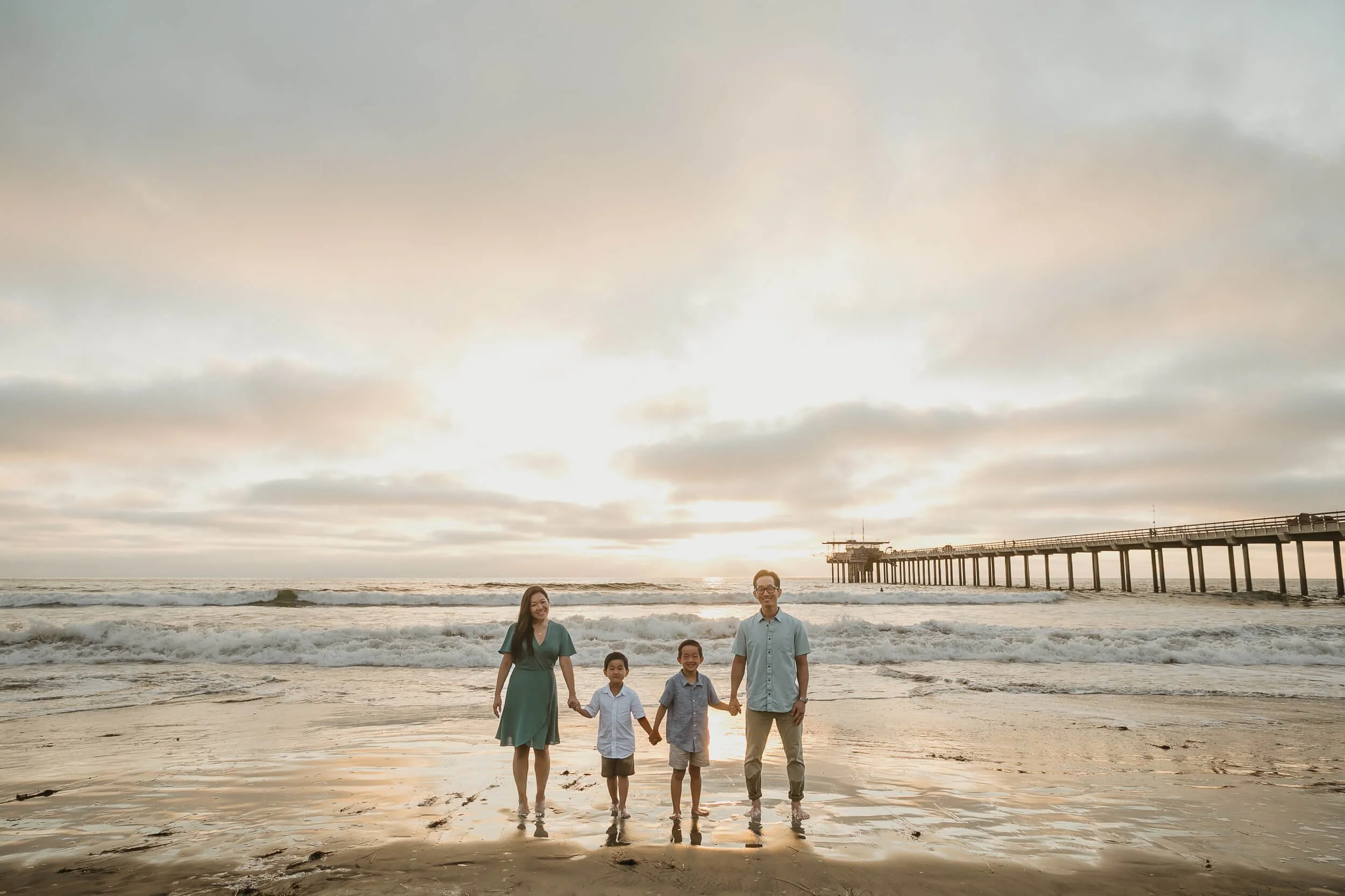 Wide photo of family of four looking at camera and holding hands during golden hour sunset at La Jolla Shores Beach
