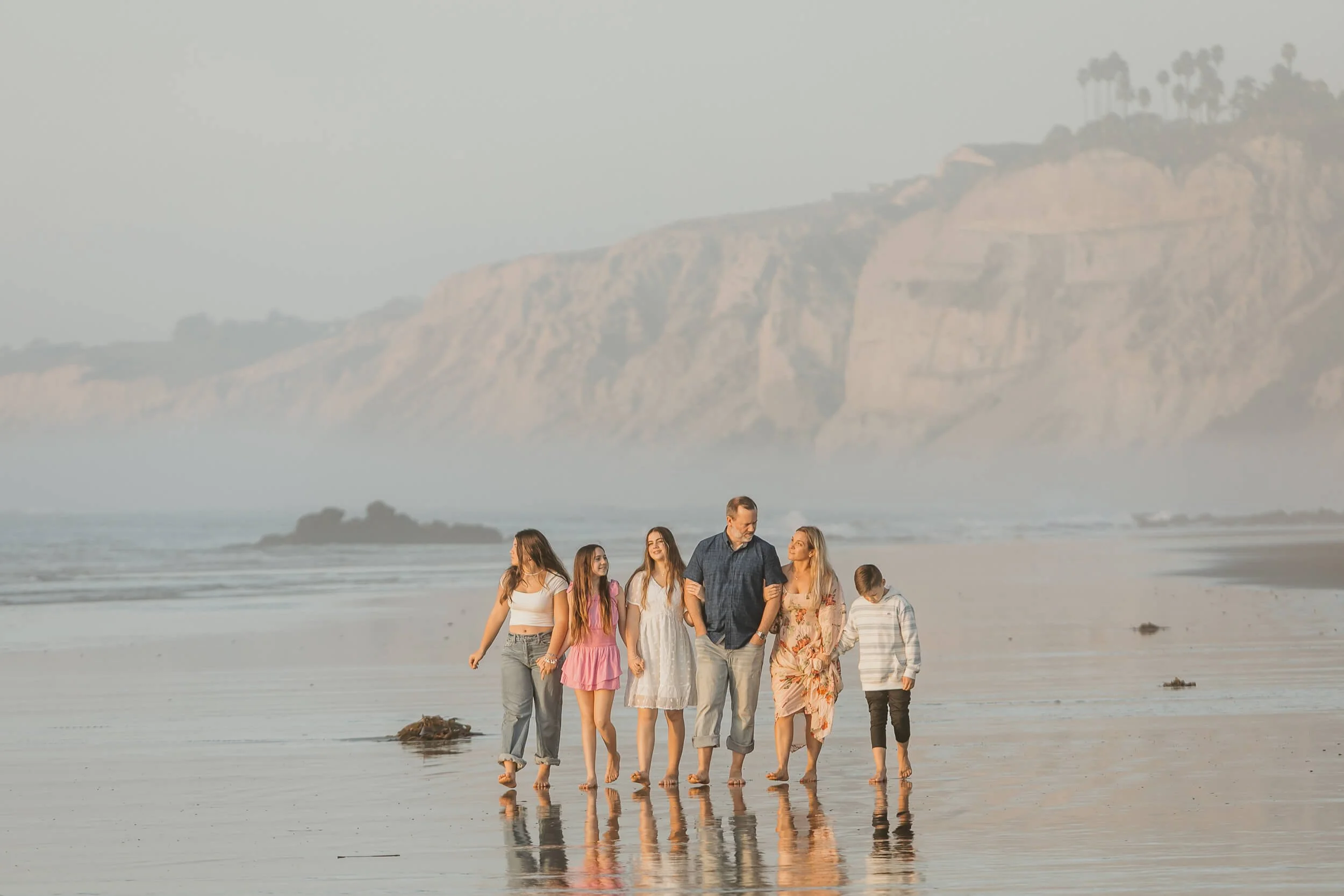 Family of six talking while holding hands and walking during low tide on family portrait session with tall Blacks Beach cliffs behind