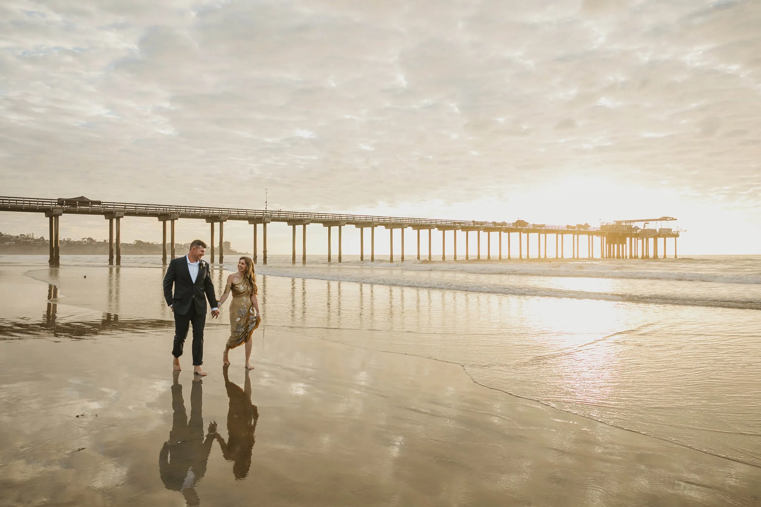 Wide shot couple looking at each other while walking on the shoreline of La Jolla Shores with Scripps Pier behind