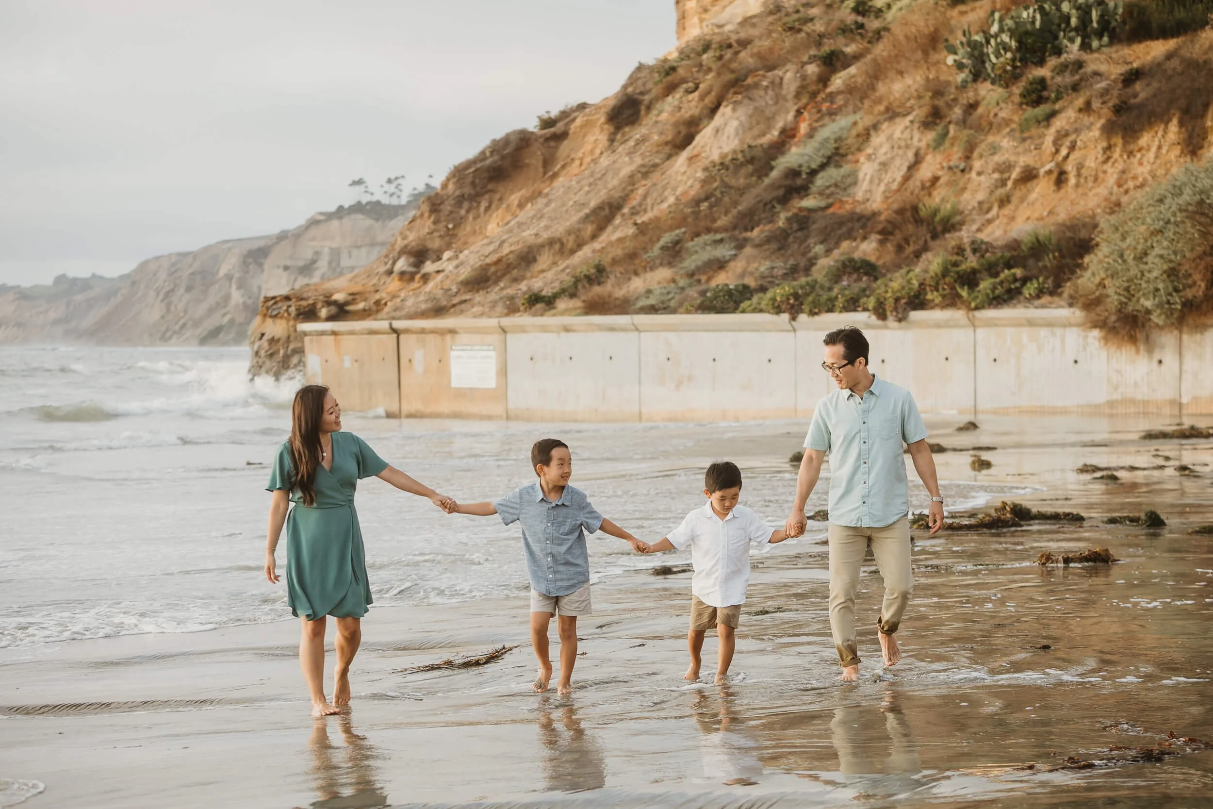 Family holding hands and walking on shoreline of Scripps La Jolla Shores Beach with Blacks Beach in the background