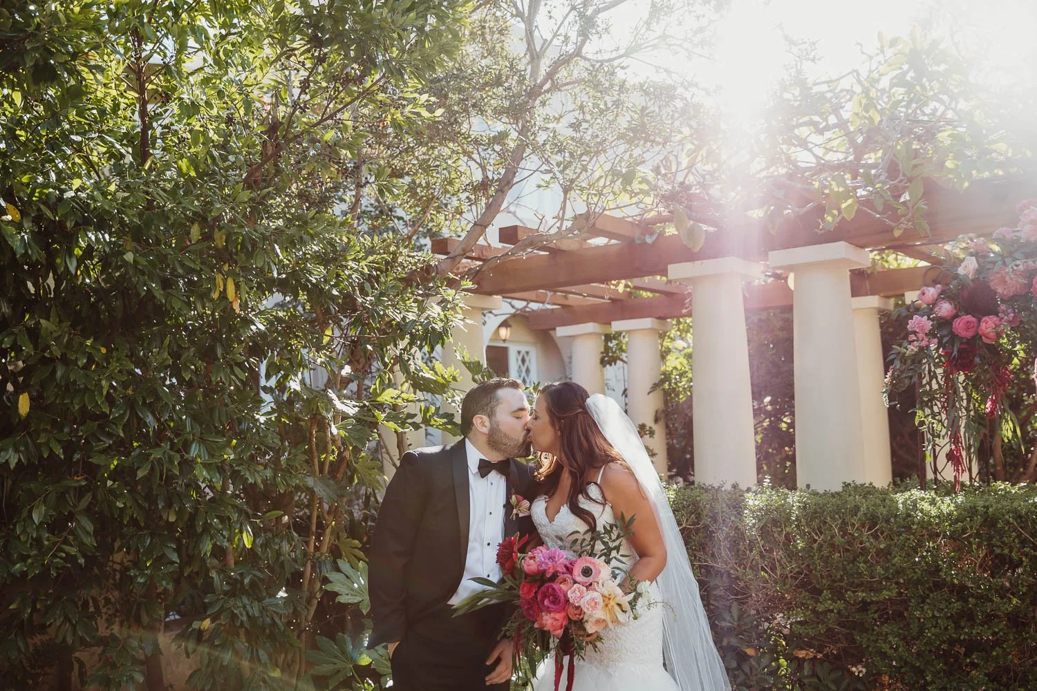 A bride and groom sharing a kiss outdoors in a garden setting with sunlight filtering through trees, the bride holding a bouquet of pink and white flowers.