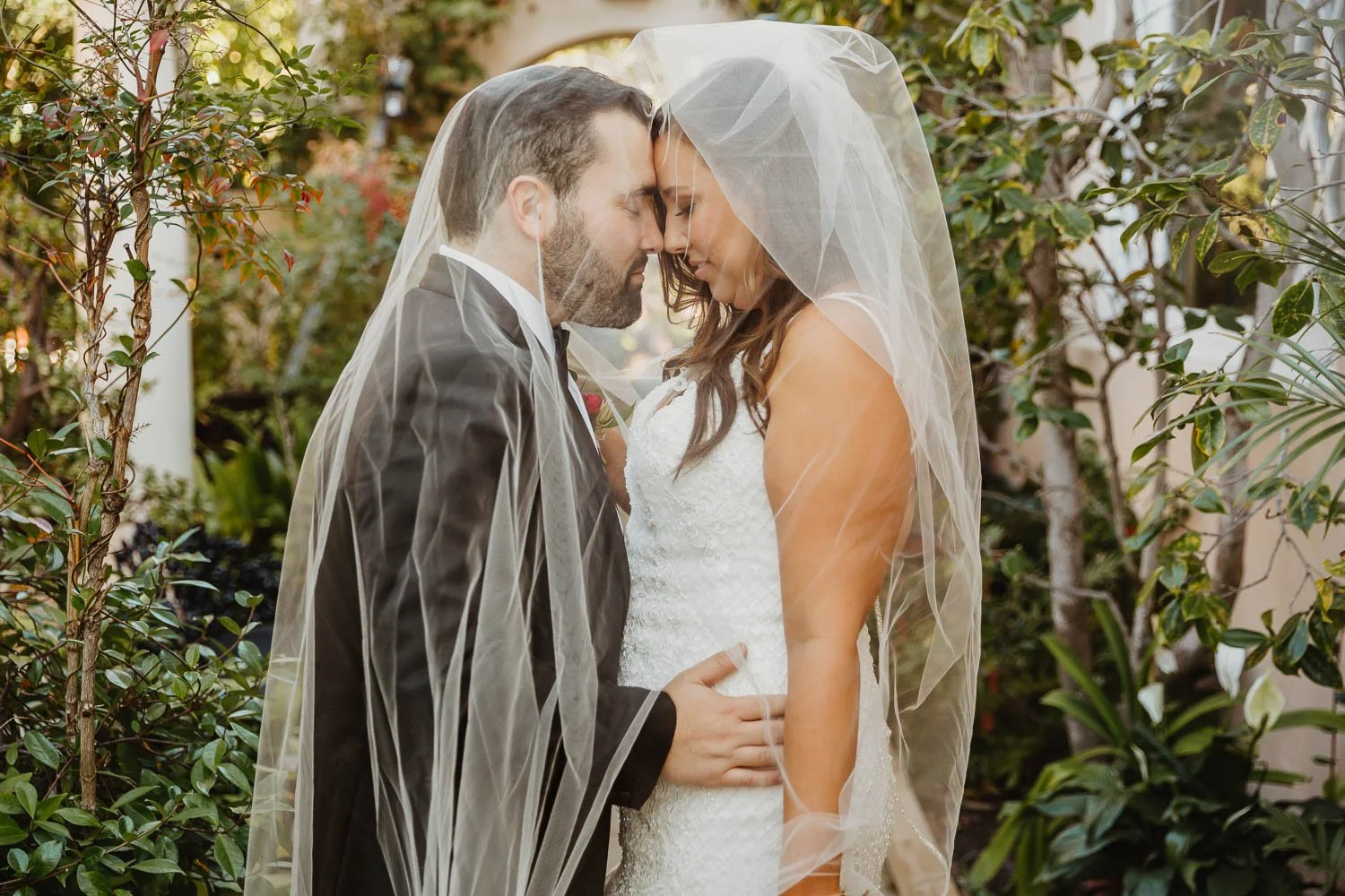A bride and groom with their foreheads touching under a veil during their wedding, surrounded by greenery.