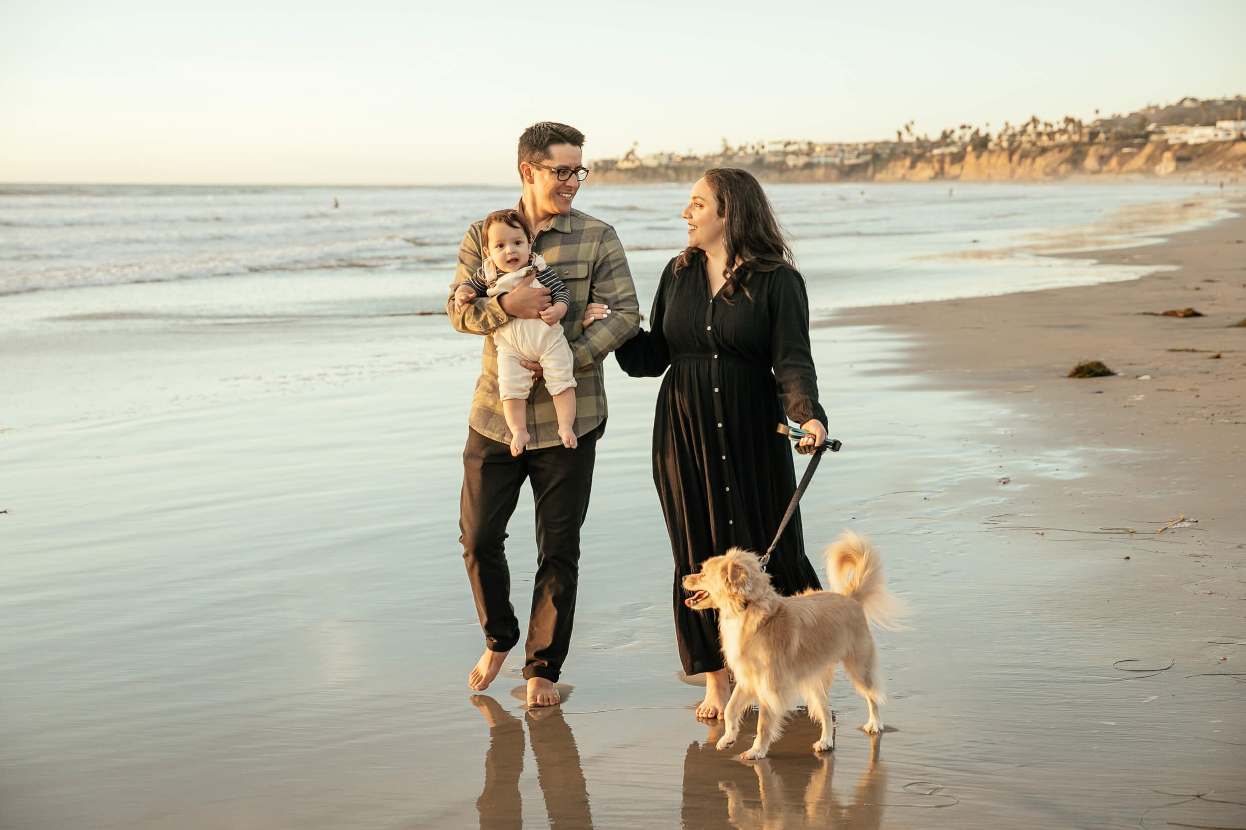 Couple walking on shoreline looking at each other holding baby and dog for Pacific Beach / Crystal Pier Family Sessions
