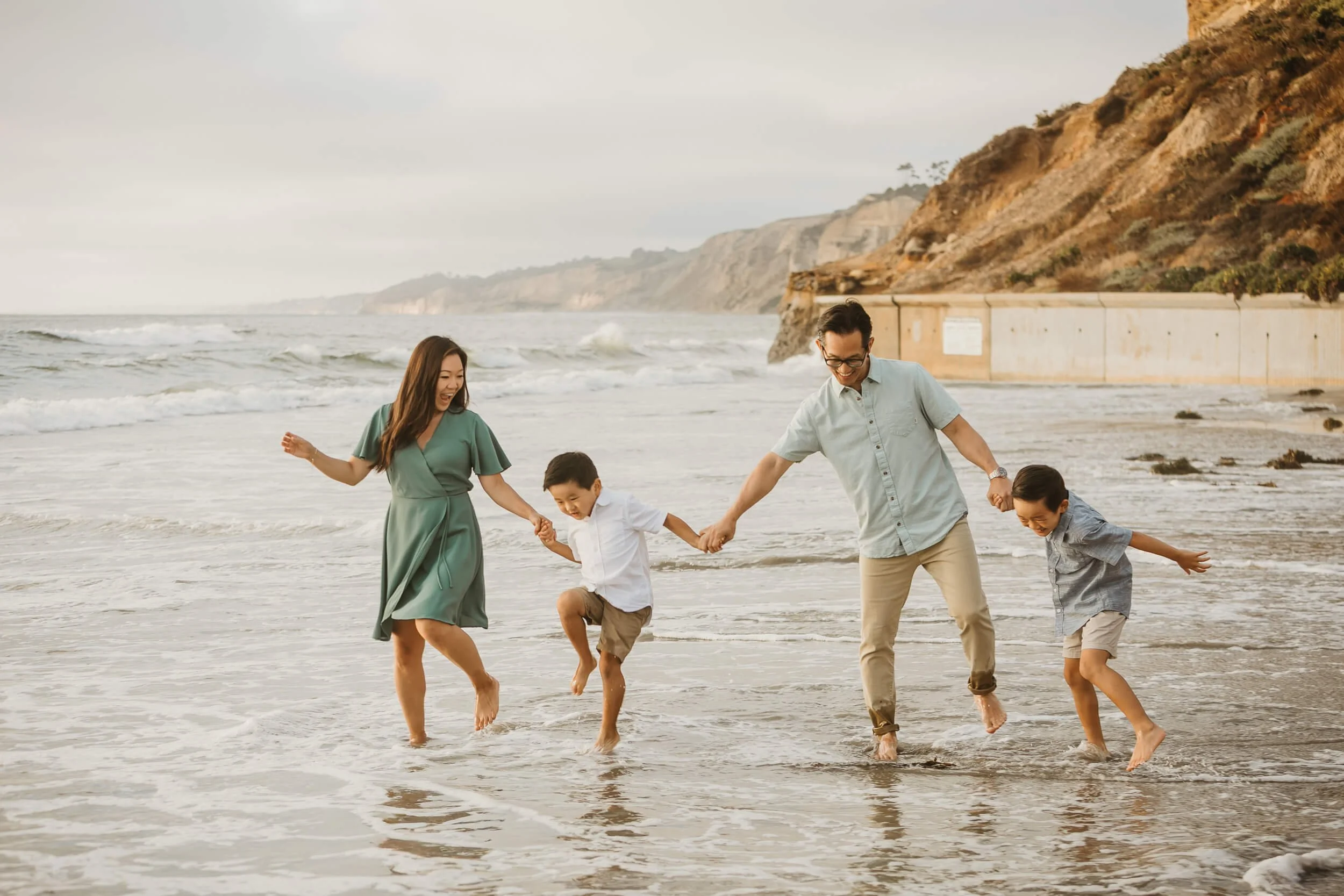 Fun moment of family of four being hit by the wave while walking holding hands during San Diego family portrait session at La Jolla Shores Beach 