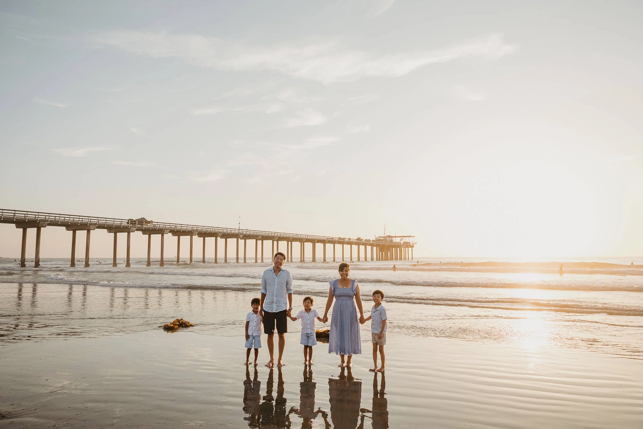 Wide shot family holding hands and looking at camera during sunset photo session at La Jolla Shores with Scripps pier behind