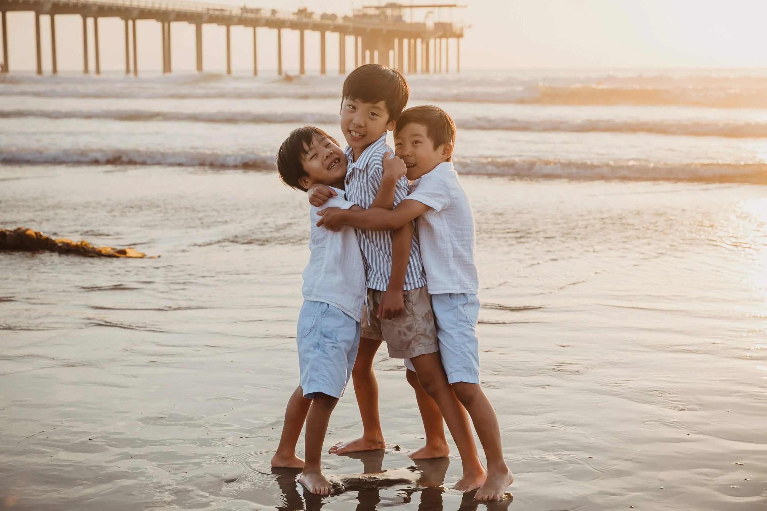 Fun photos of three boys bothers hugging each other with sunset and Scripps pier behind 