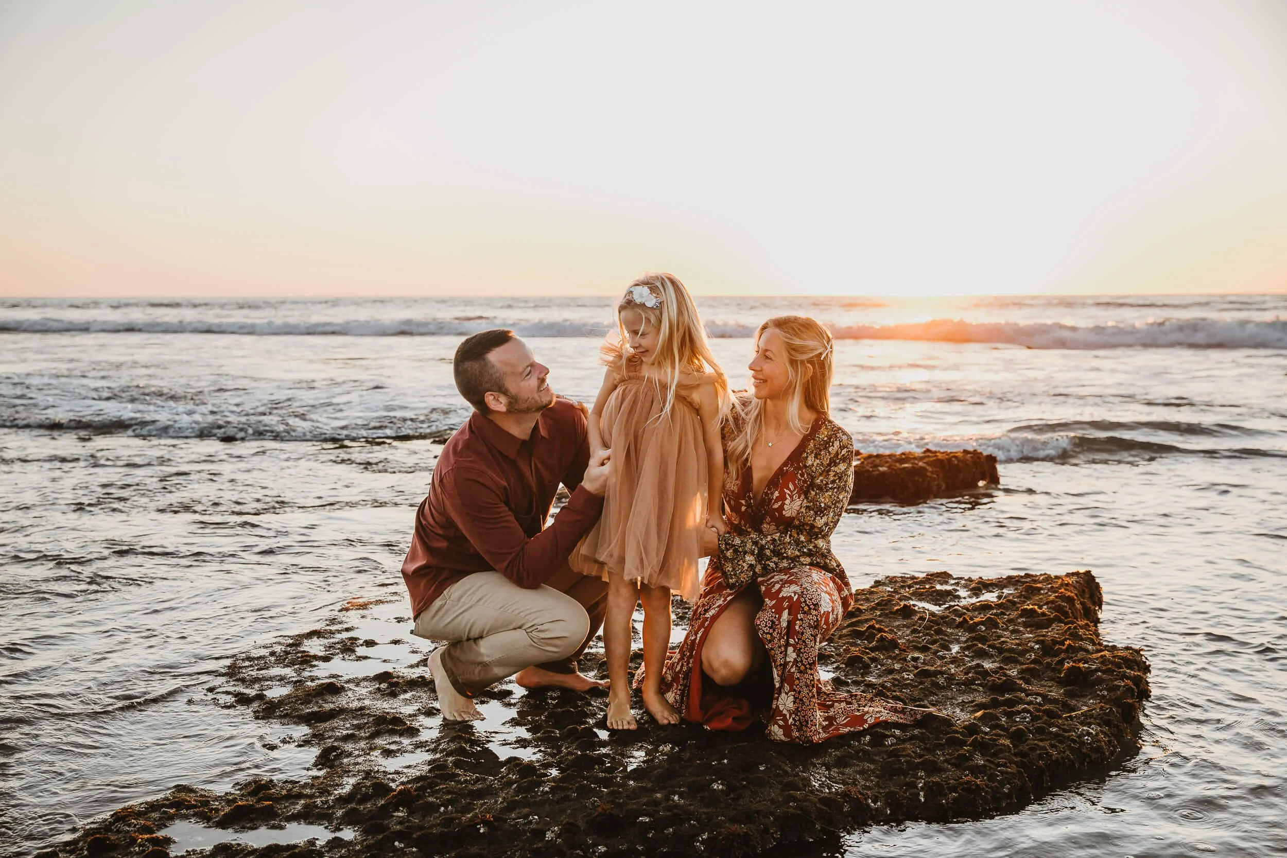 A family of three on a rocky beach during sunset, with the father holding his young daughter and the mother kneeling beside them, all smiling.