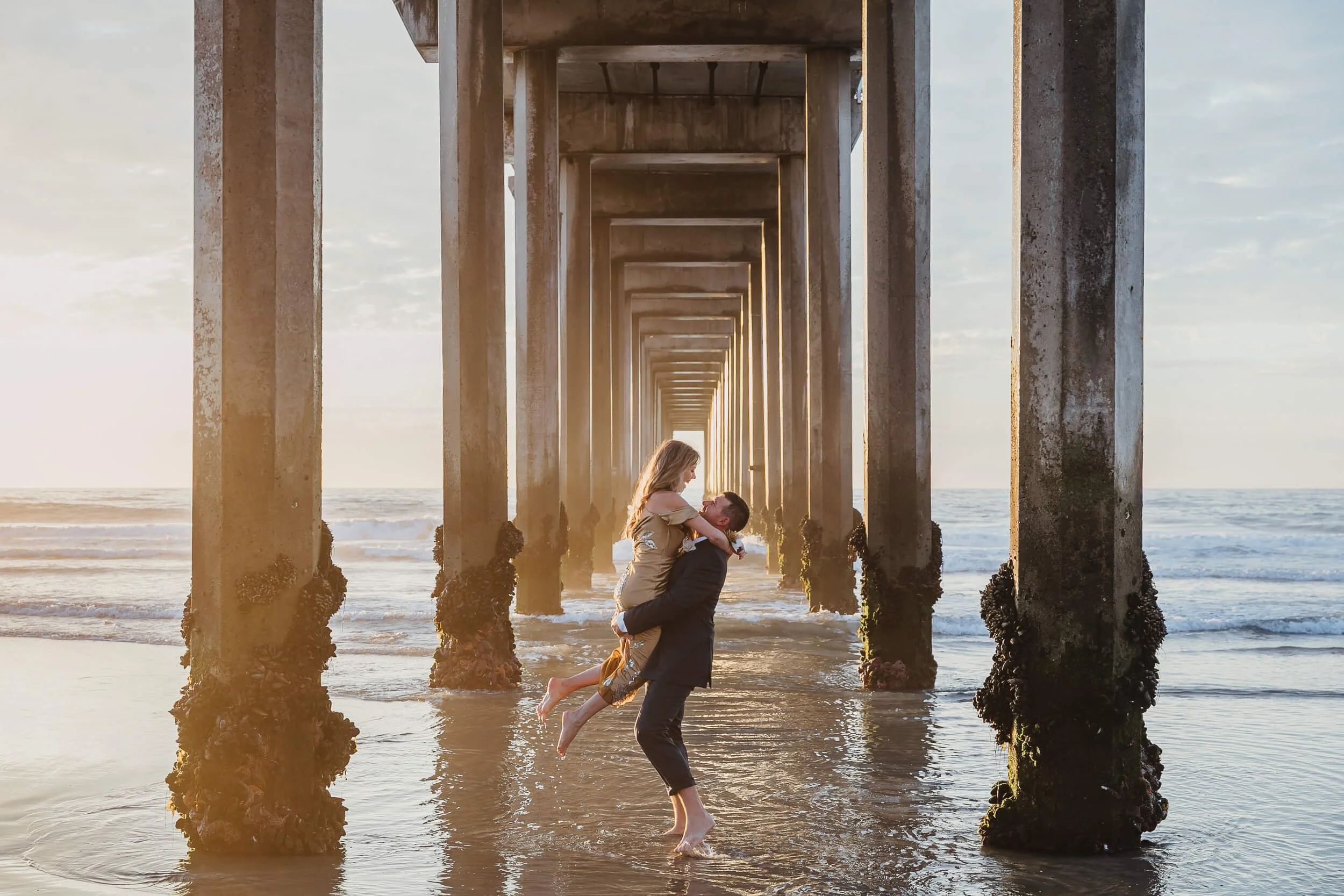 Man lifting woman under the Scripps Pier in La Jolla symmetric romantic portrait during San Diego mini session