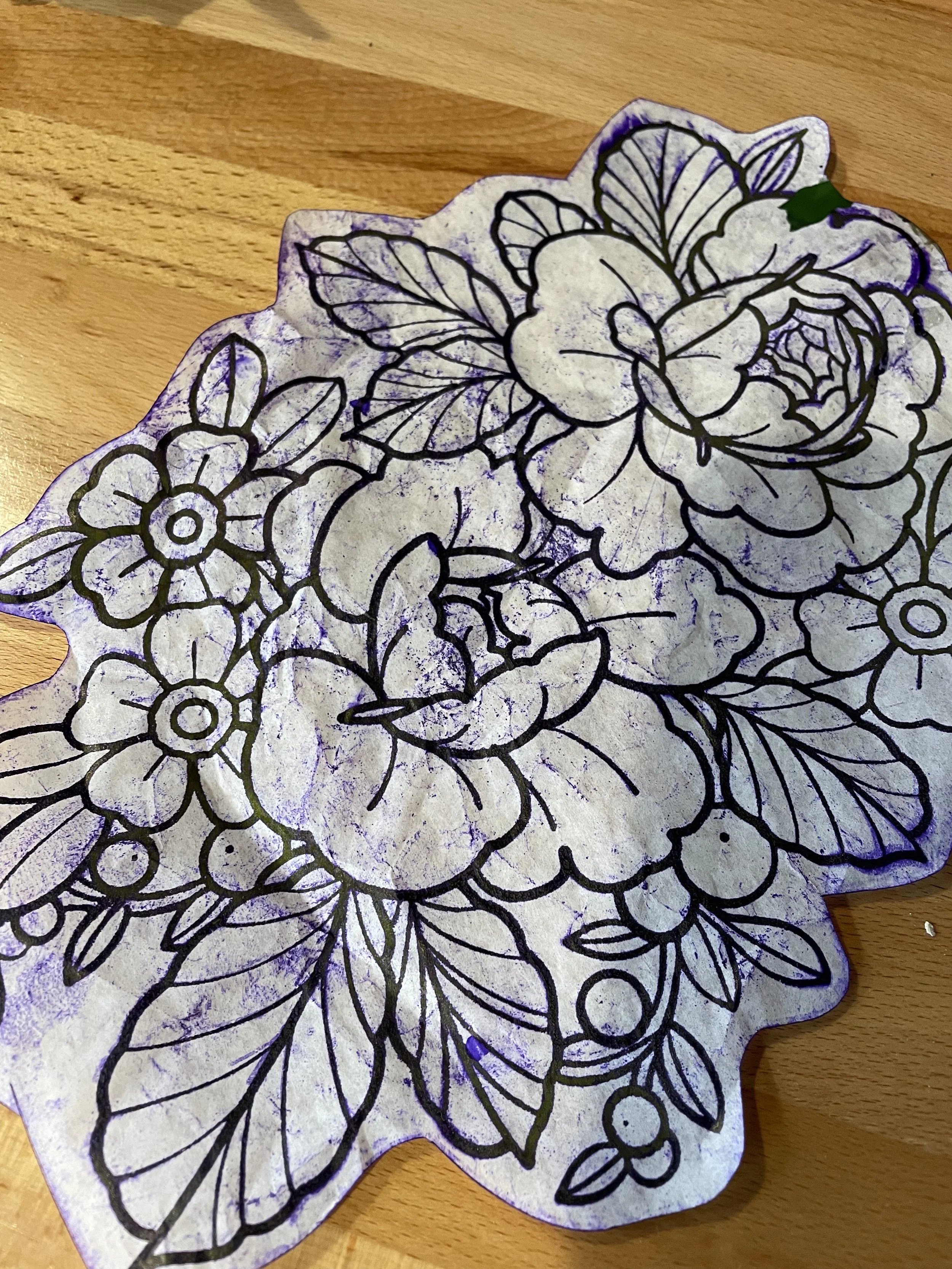 Colored paper with floral and leaf designs outlined in black, partially cut out, on a wooden surface.