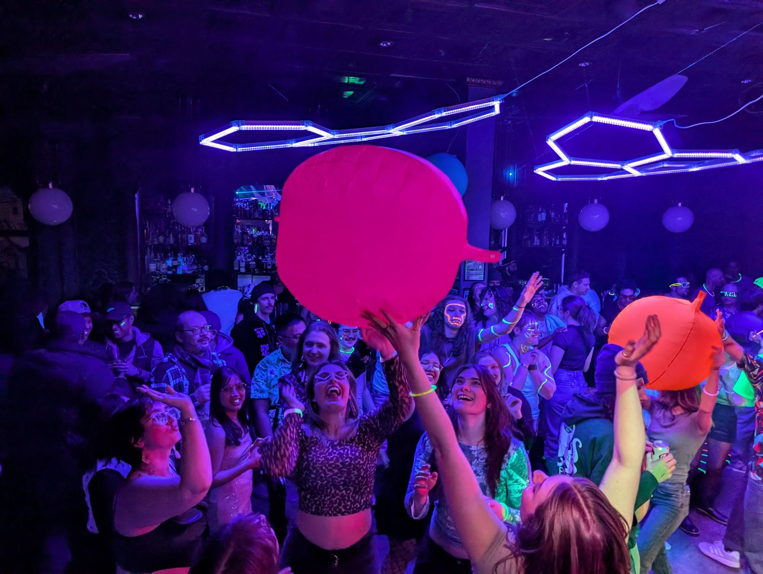 A lively crowd at a neon-lit party, with people dancing and smiling, holding glow-in-the-dark umbrellas, in a dark room with purple and blue neon lighting.