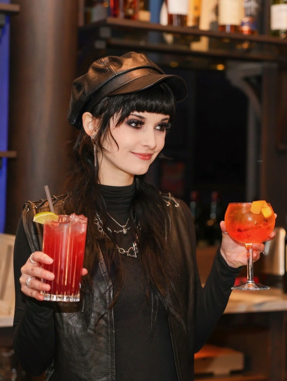 A woman with dark hair and makeup, wearing a black leather cap and black clothing, holding two cocktails in a bar or restaurant.