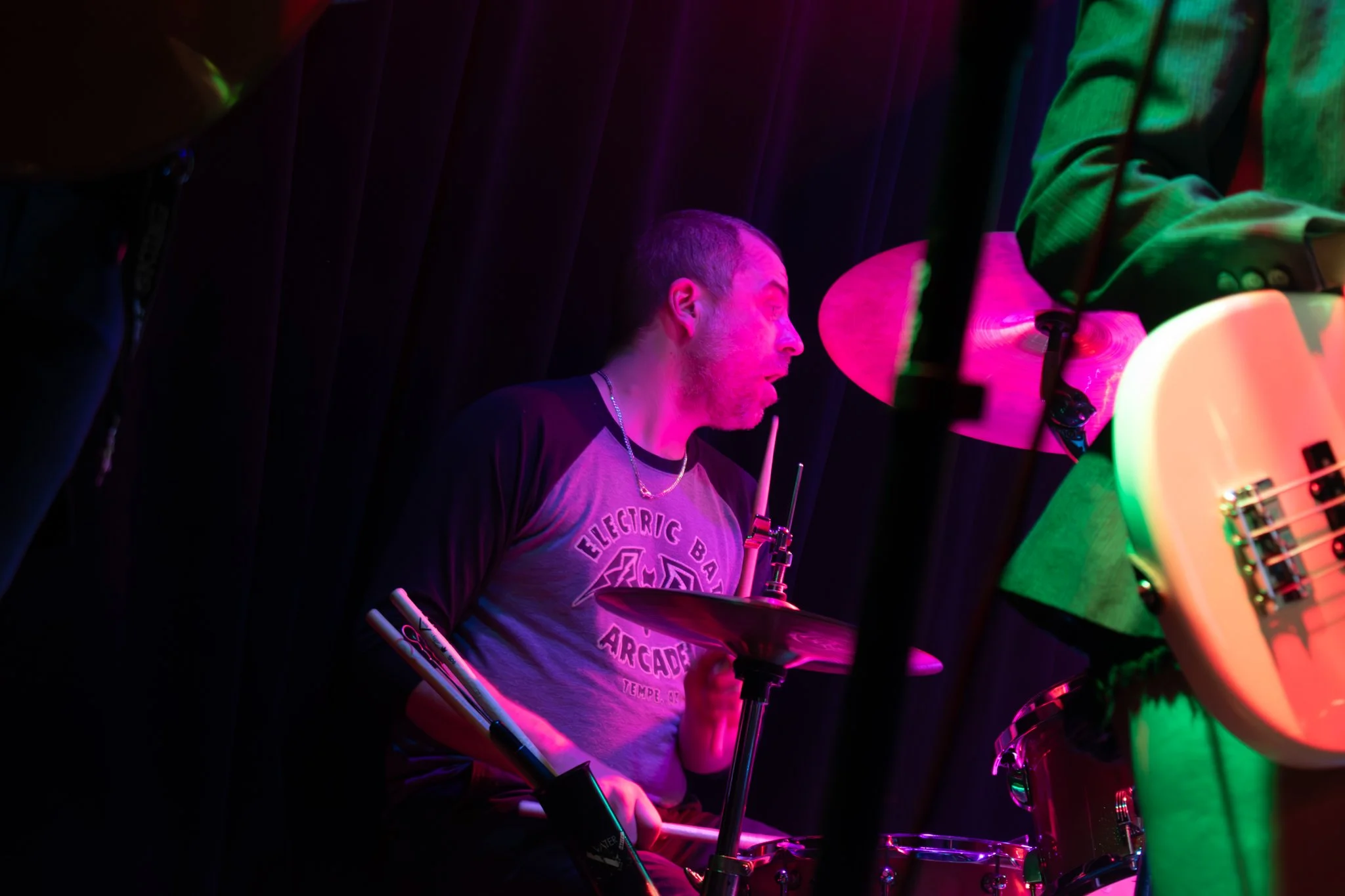 A man playing drums on stage, illuminated by colorful stage lights with dark curtains in the background.