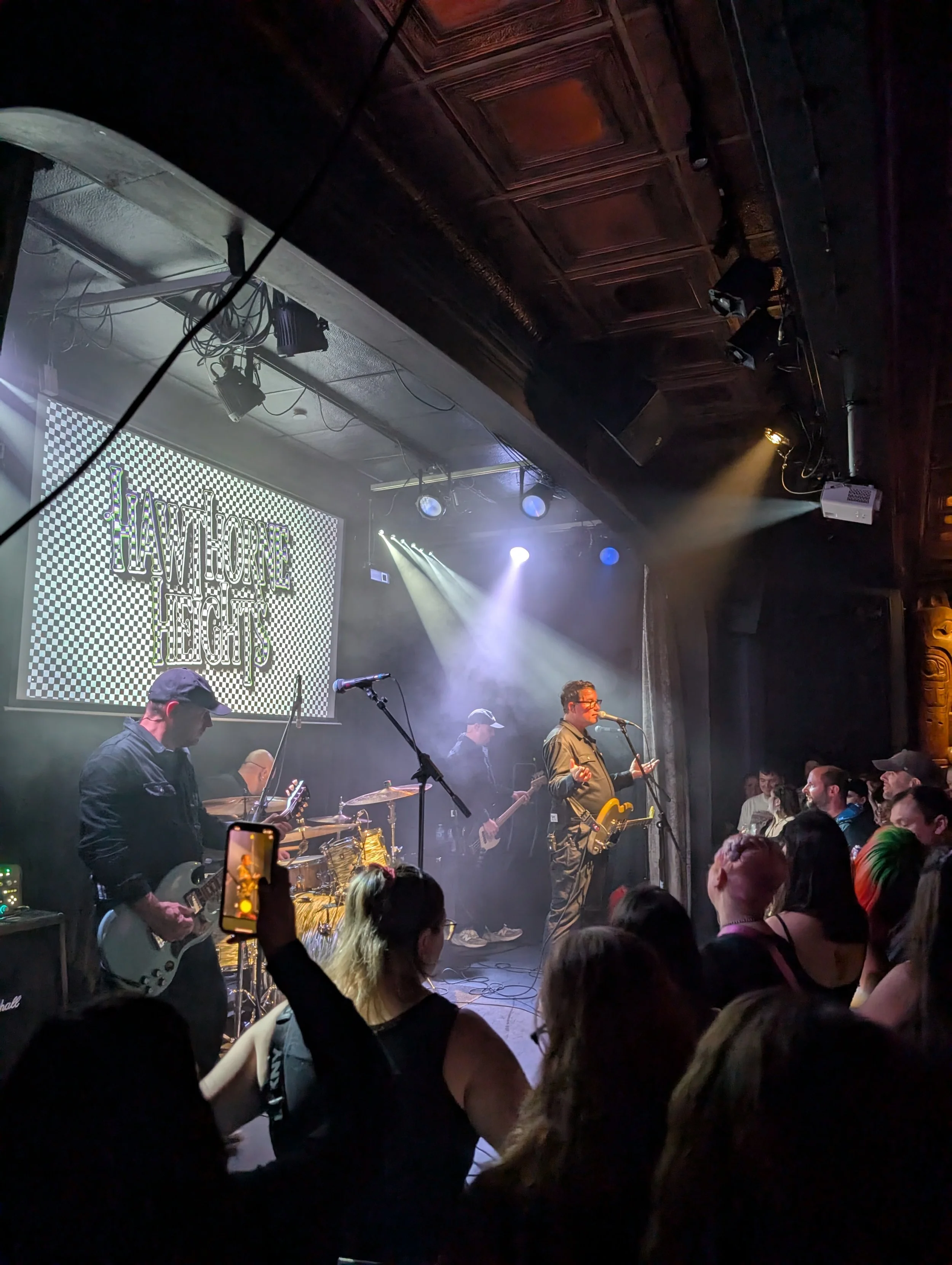 A live music concert taking place in a dimly lit venue with a band performing on stage. The band includes a guitarist and a vocalist with glasses. The audience is engaged, with some taking photos and videos. There is a large LED screen in the backgro