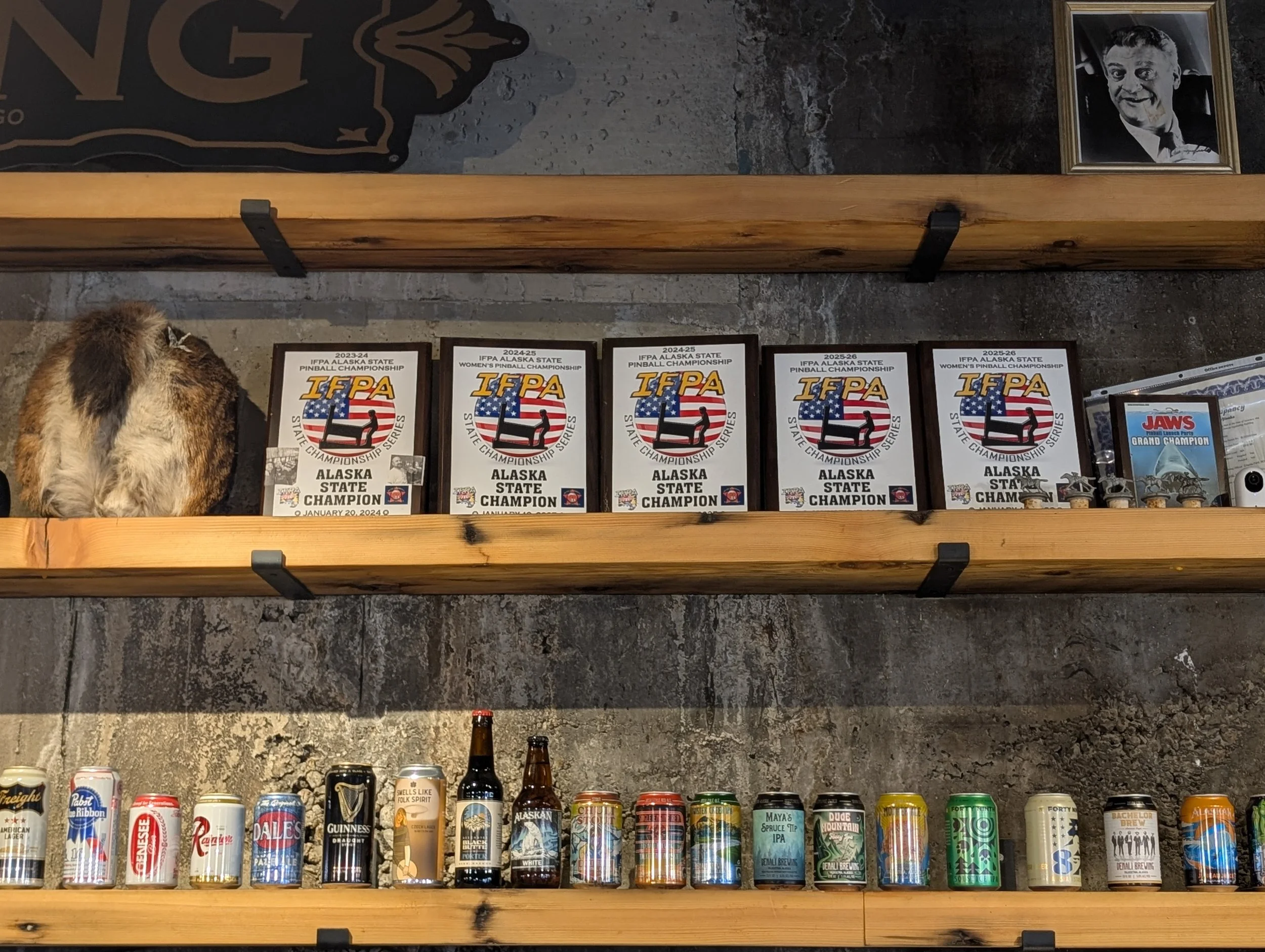 Shelf with multiple award plaques for the Alaska State Pinball Championship from 2023 to 2025, and a black and white framed photo of a smiling man with gray hair in a tuxedo.