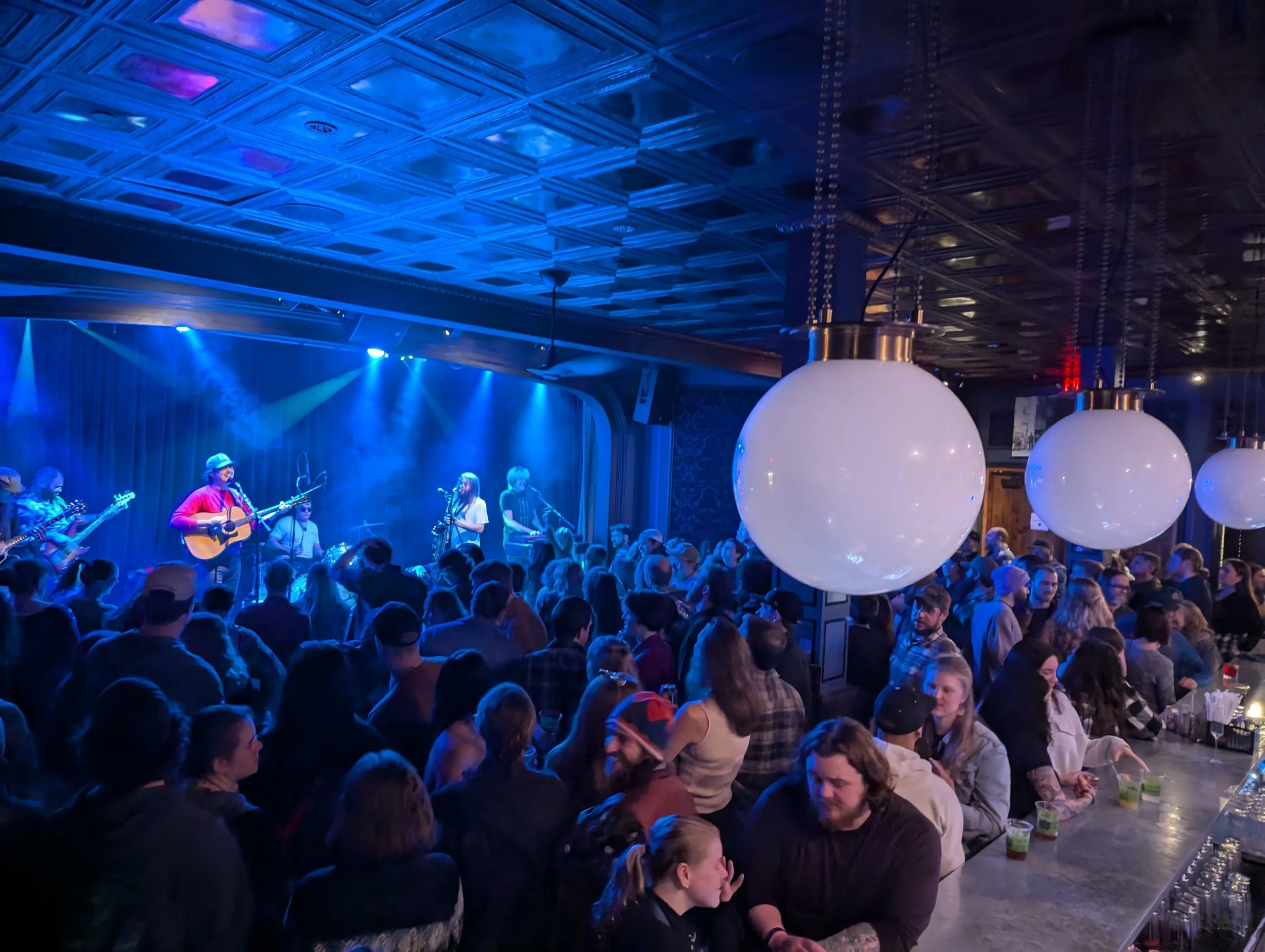 People gathered in a dimly lit venue watching a band perform on stage illuminated with blue and purple lighting, with large white spherical lights hanging from the ceiling.