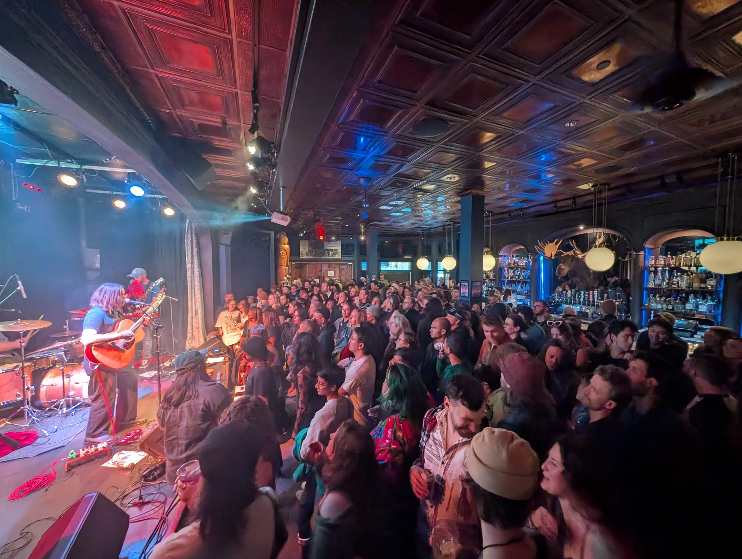 Crowd of people at a live music performance inside a dimly lit bar or club. Musicians playing guitar on stage with colorful stage lights overhead and bar area visible in the background.