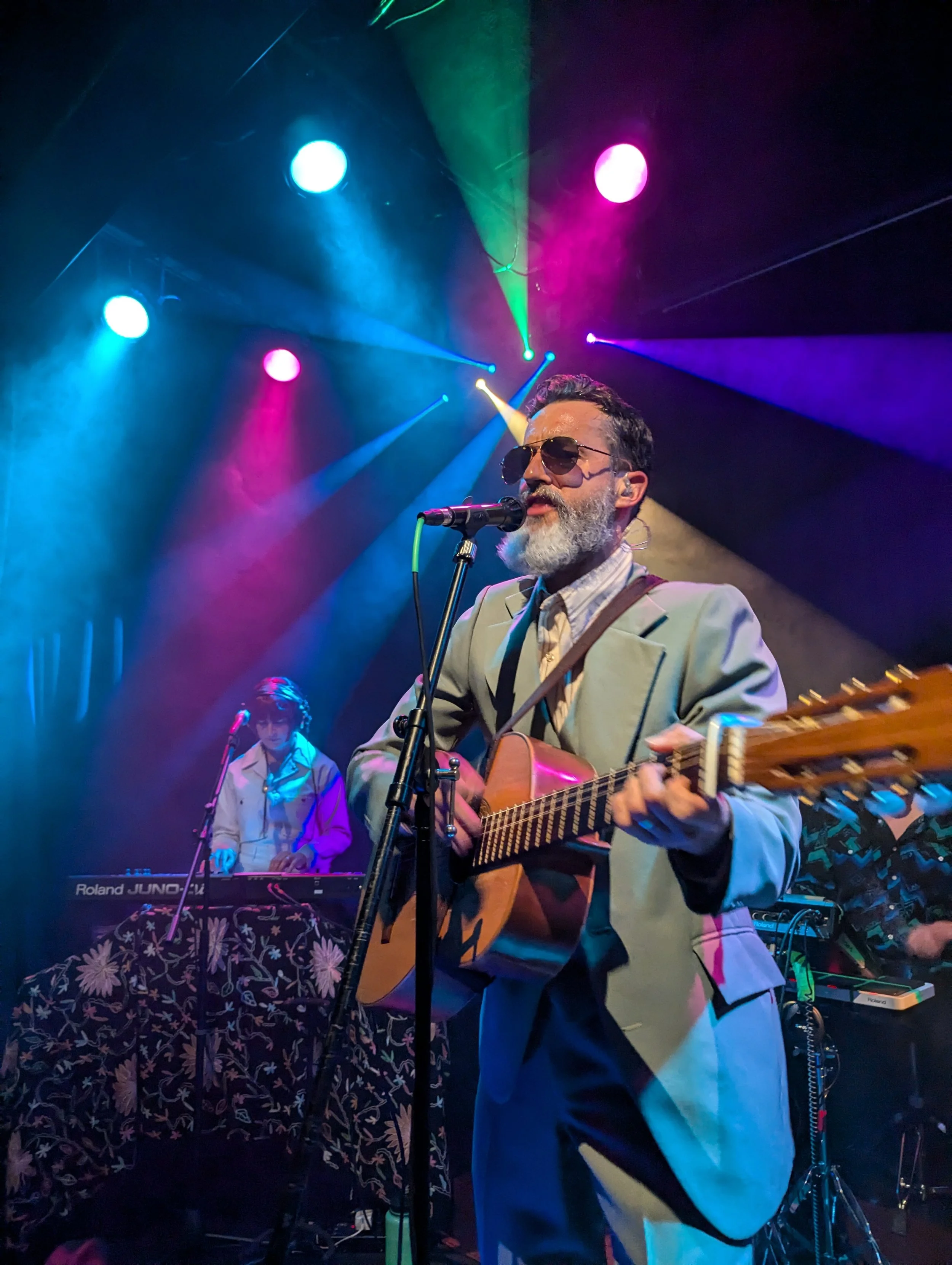 A male musician with a beard, wearing sunglasses and a light-colored blazer, is singing and playing an acoustic guitar on stage during a live performance. The stage is lit with vibrant, colorful lights including blue, pink, green, and purple. In the 