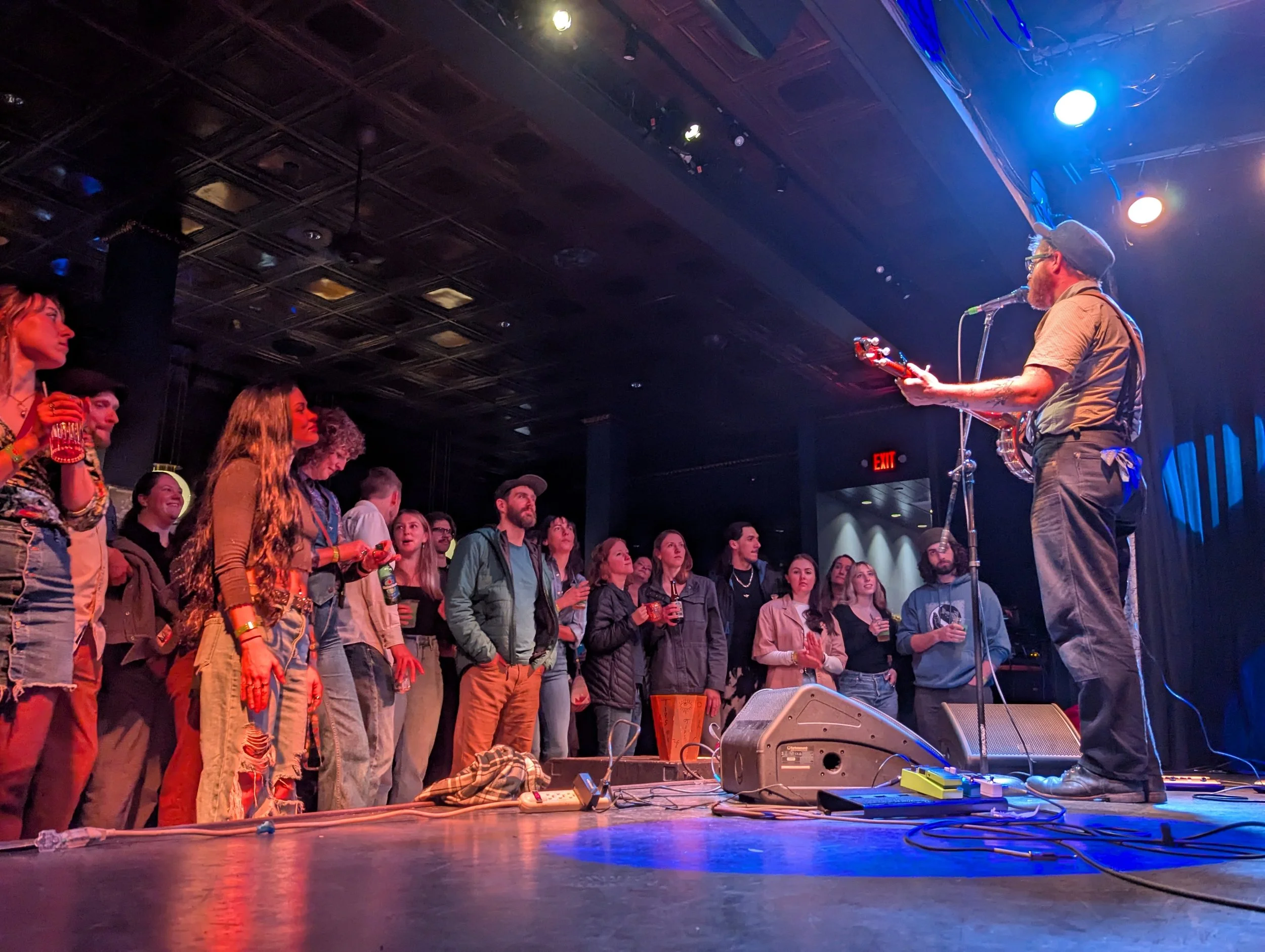 A musician with a beard and glasses plays guitar and sings into a microphone on stage in front of an audience. The audience, composed of diverse people, is attentively watching the performance in a dimly lit venue with blue and orange stage lighting.