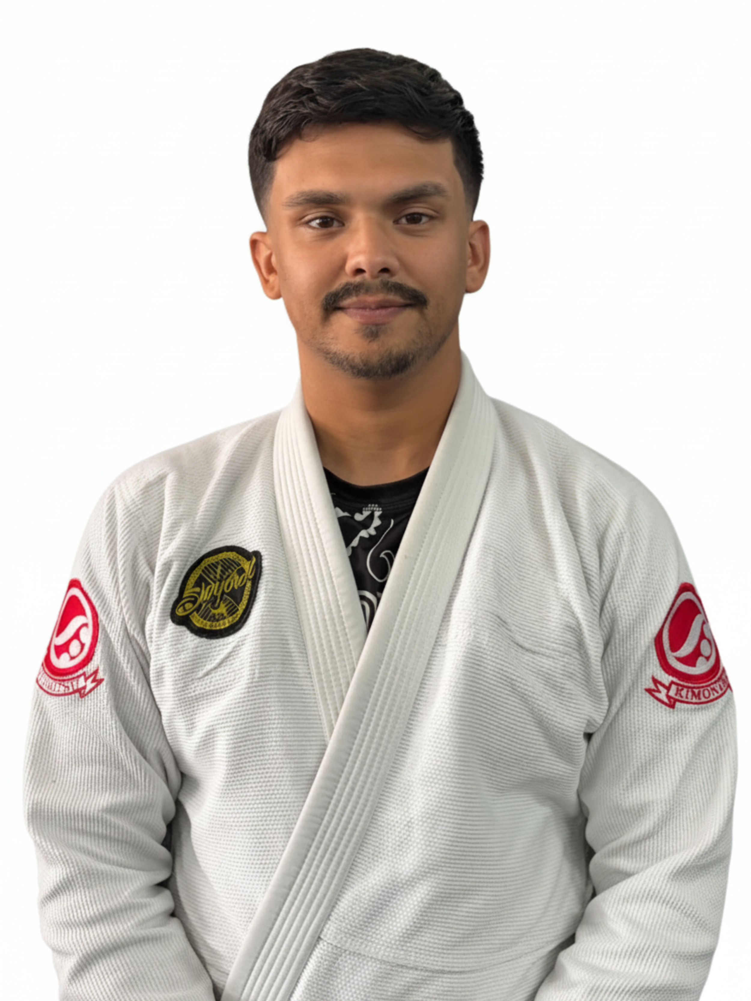 A young man with dark hair and a mustache wearing a white Brazilian Jiu-Jitsu gi with various patches, including a yellow and black seal patch on his left chest and a red patch on his right sleeve, standing against a plain white background.