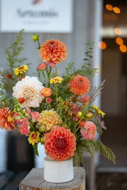 Colorful flower bouquet in a white vase on a wooden pedestal, with orange, yellow, pink, and white flowers and greenery.
