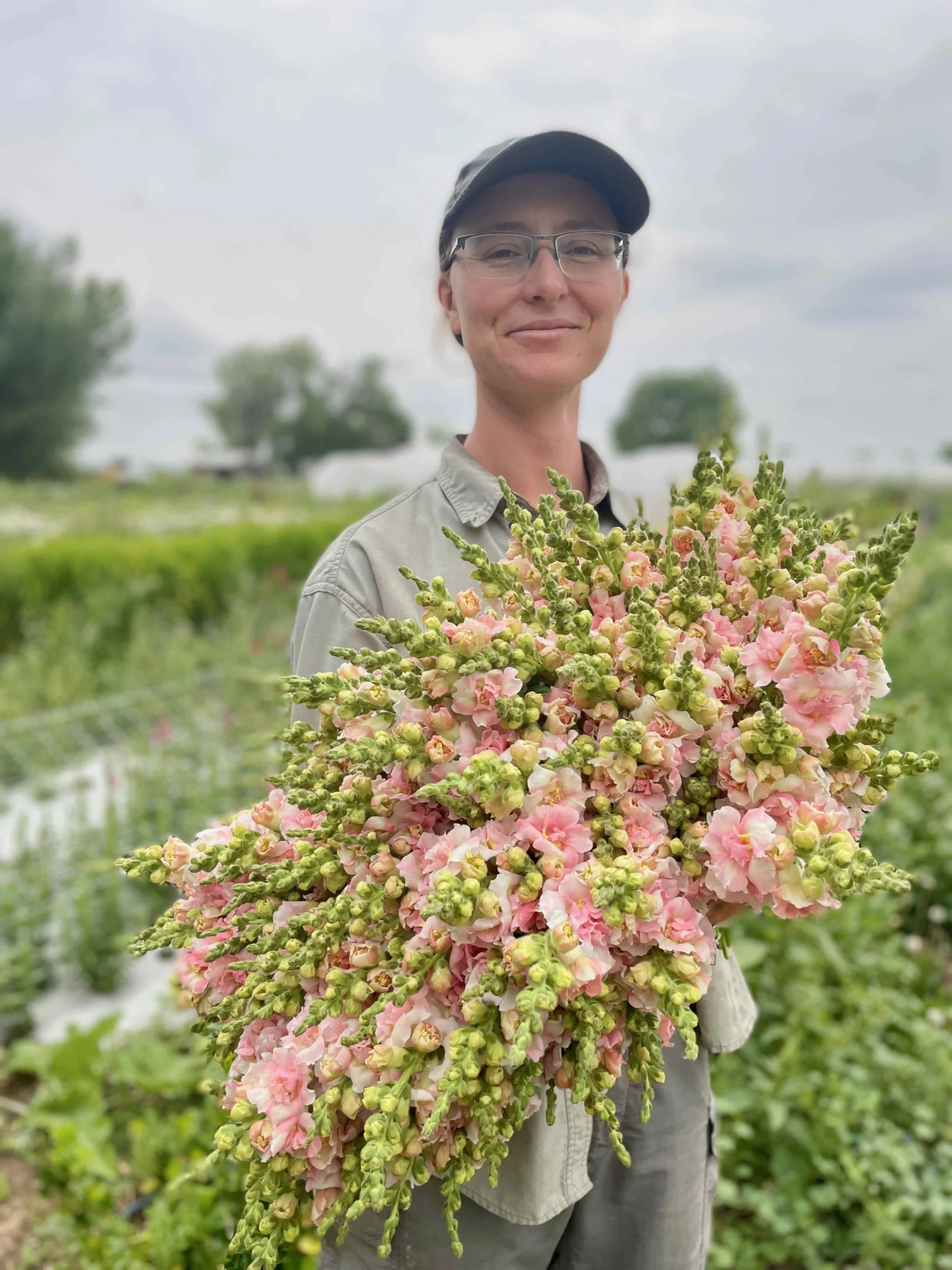 Woman holding a large bunch of pink and green snapdragons in a field, wearing glasses, a khaki shirt, and a black cap, with a cloudy sky overhead.