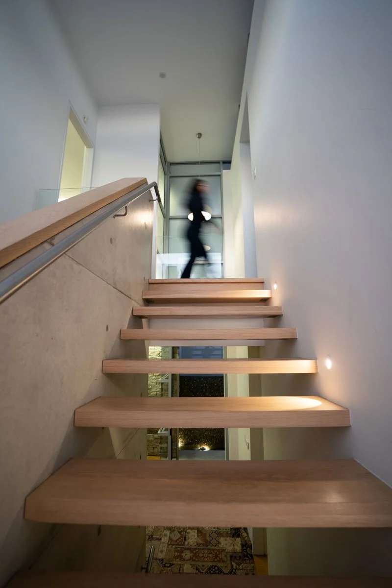 An upward view of a staircase inside a modern building, with a person blurred as they walk up the stairs, and illuminated small lights on the right wall.