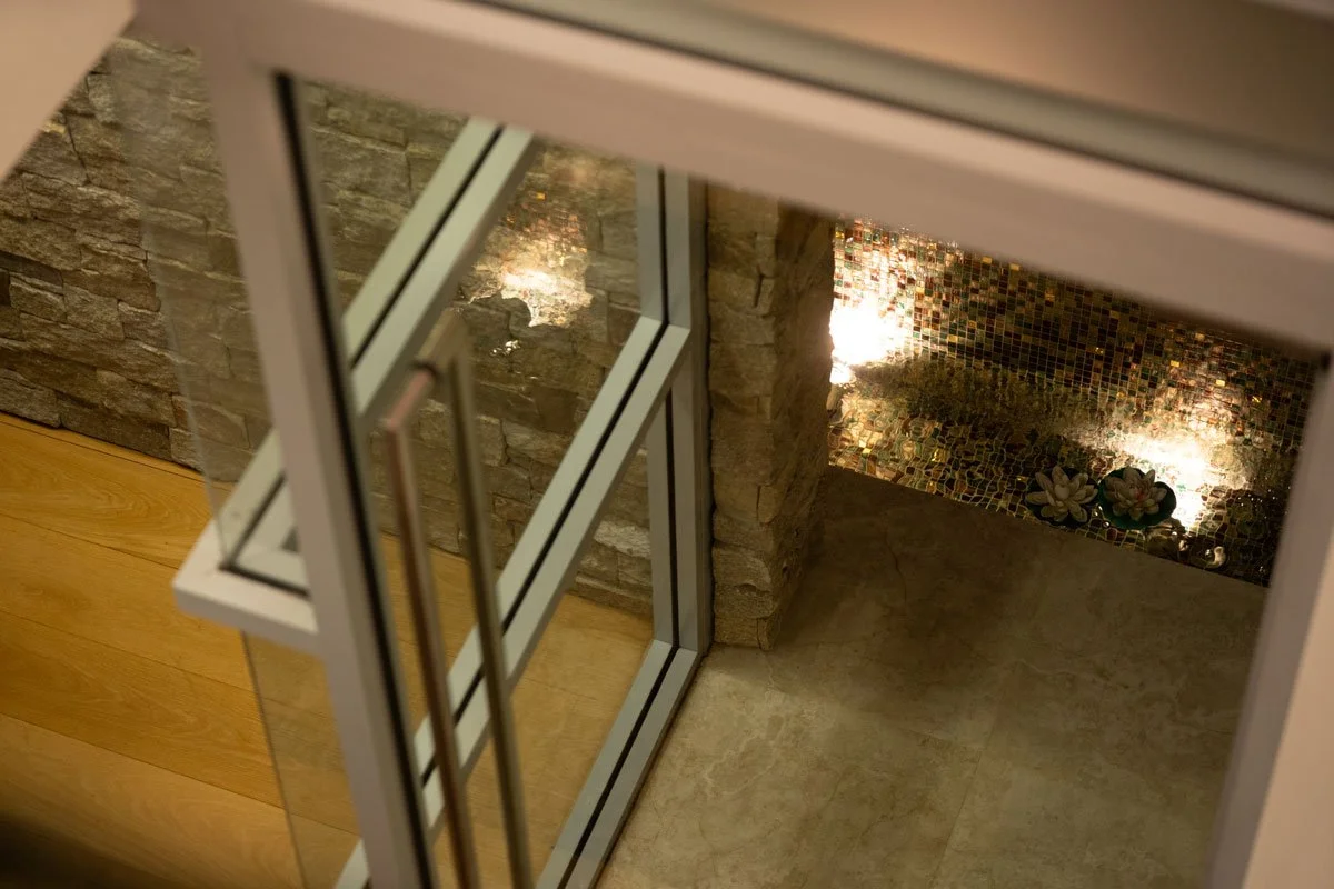 View through a glass door showing a stone wall, a wall with small decorative tiles, and some potted plants illuminated by lights.