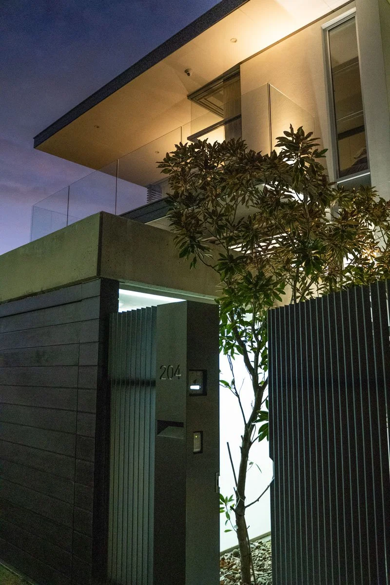 Modern house exterior at dusk with a gate and a tree illuminated.